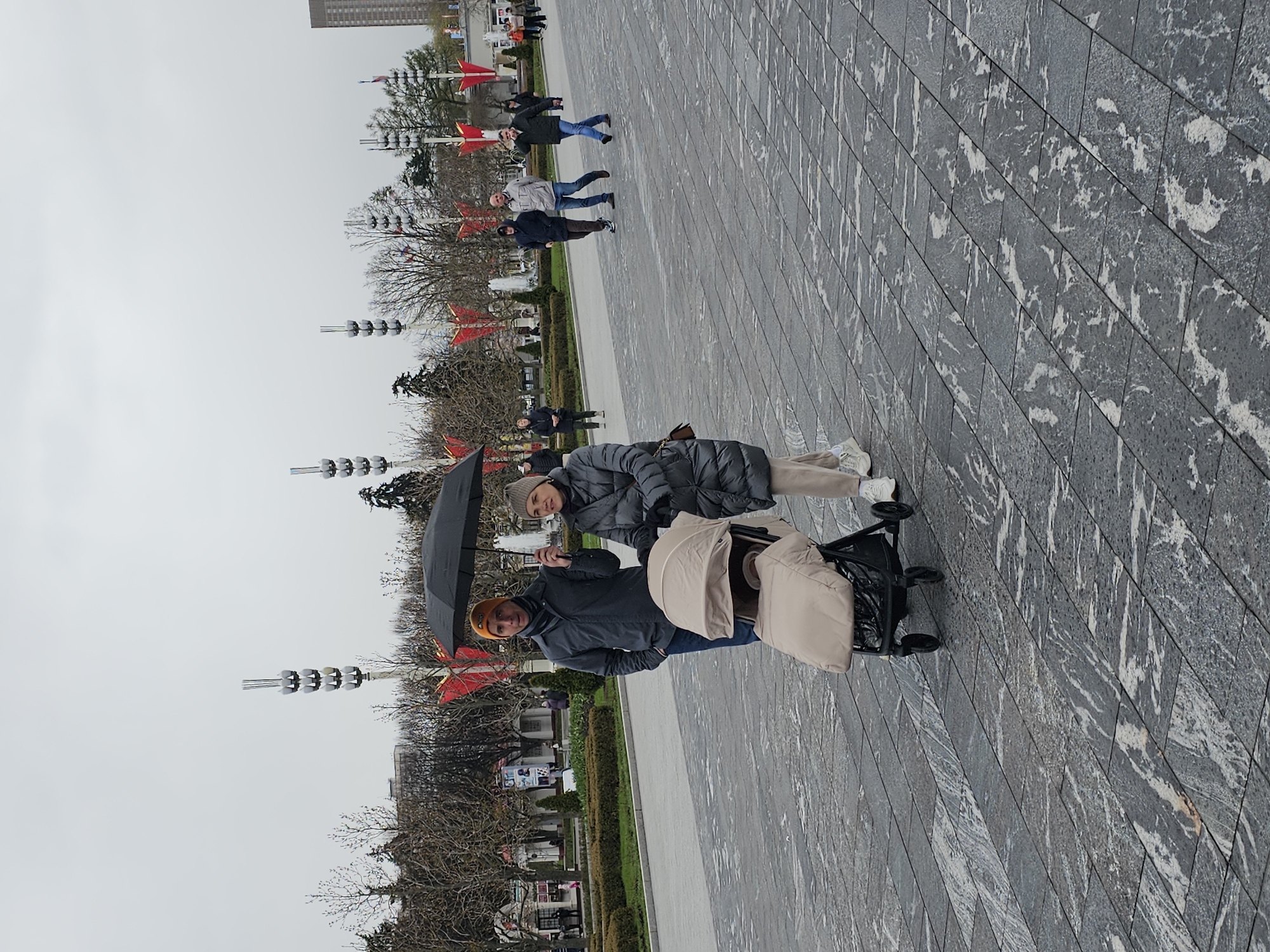 This image captures an outdoor scene in a public square or wide promenade in Khimki, Russia, under an overcast sky. In the foreground, two adults are walking side-by-side. One individual, wearing a dark jacket and an orange beanie, holds a black umbrella, indicating possible rain or a readiness for it. The other adult, dressed in a grey puffy jacket and a light-colored beanie, is pushing a beige baby stroller with a black frame.
The ground beneath them is covered with large, rectangular dark grey tiles featuring prominent white, marble-like veining, creating a distinctive pattern. In the midground, several other people are visible walking, dressed in casual outerwear suitable for cool weather. The area is landscaped with bare deciduous trees, suggesting late winter or early spring, and some evergreen shrubs. Stylized lamp posts with multiple circular light fixtures are spaced throughout the area, alongside poles adorned with red, star-shaped decorations. In the distant background, a tall, light-colored building rises against the uniformly grey sky. No explicit text is clearly visible. The overall mood is calm, depicting everyday activities in a public urban space.