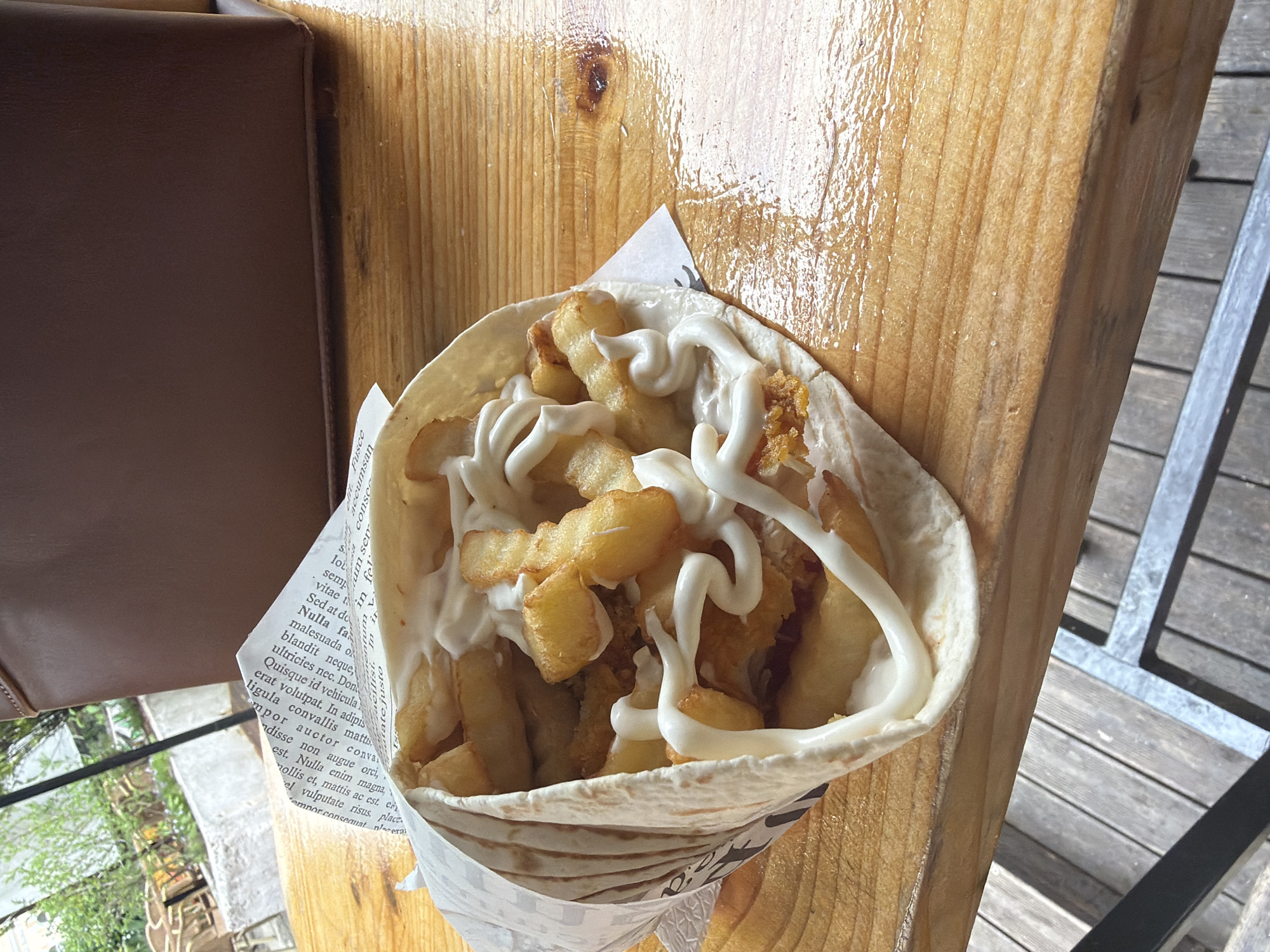 A close-up shot depicts a handheld wrap filled with french fries and a white sauce, possibly mayonnaise. The wrap is presented on a wooden surface with visible grain and an apparent glossy finish, suggesting a recently cleaned or wet surface. The wrap is partially contained within a wrapper patterned with printed text. To the left of the wrap, a portion of a brown cushioned surface is visible, and to the right, a section of a metal and wood slatted structure can be seen, suggesting an outdoor seating area. The environment implies the image was taken at a food establishment, potentially in Vratsa, Bulgaria.