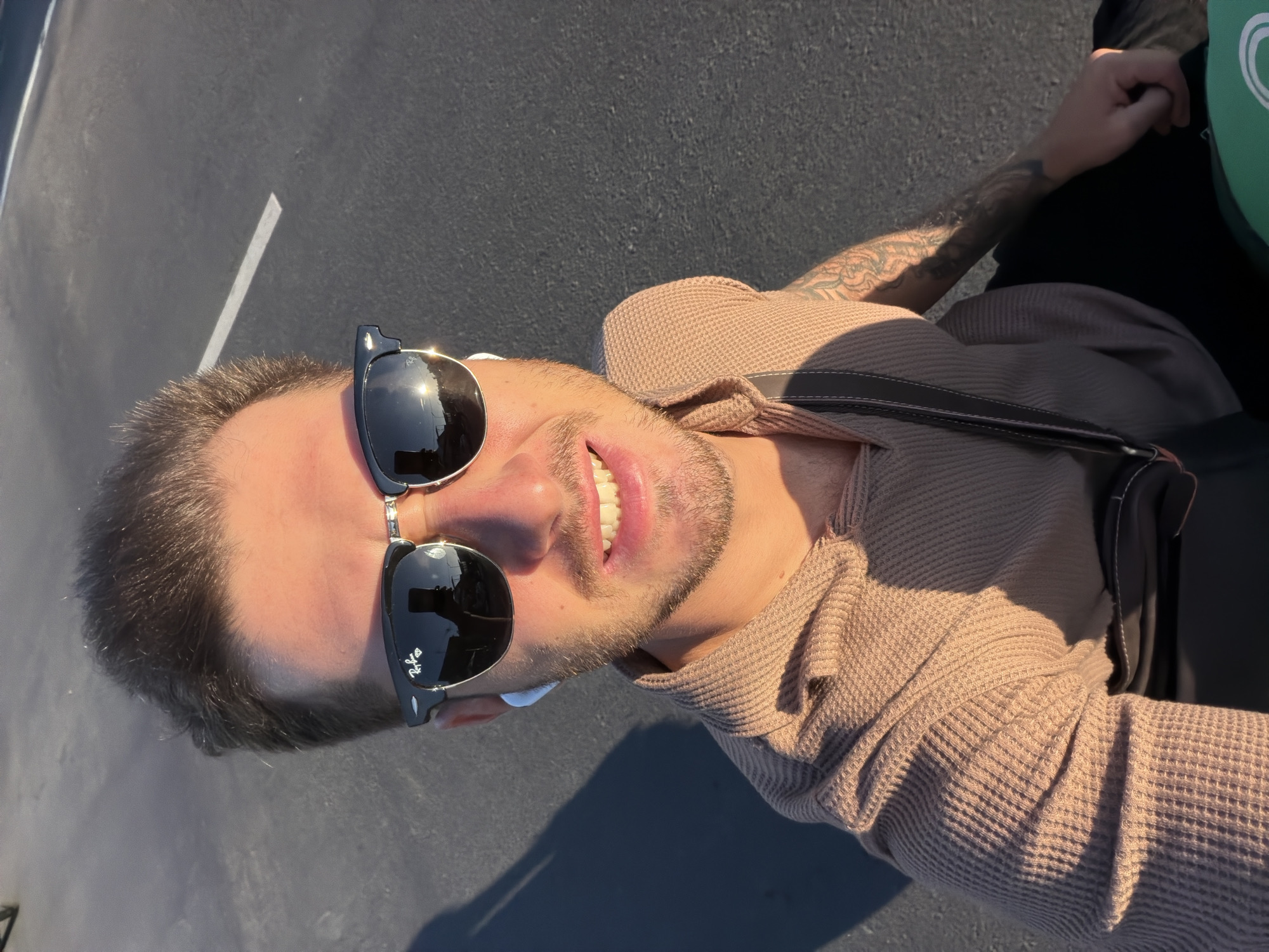 A man is captured in a bright, sunlit selfie, likely taken outdoors in North Kuta, Indonesia. He is smiling widely, revealing his teeth, and looks directly at the camera. He has short dark hair and a short beard or stubble. He is wearing black sunglasses, with the "Ray-Ban" logo visible on the right lens, and small white earbuds are visible in his ears. His attire consists of a light brown waffle-knit long-sleeved shirt, and a dark bag strap crosses his chest. Tattoos are visible on his right arm. The background shows a dark gray paved surface, resembling asphalt or a road, with a distinct white painted line visible on the left. The strong, direct sunlight indicates a clear daytime environment and casts prominent shadows on the ground. The man's cheerful expression suggests a positive mood.