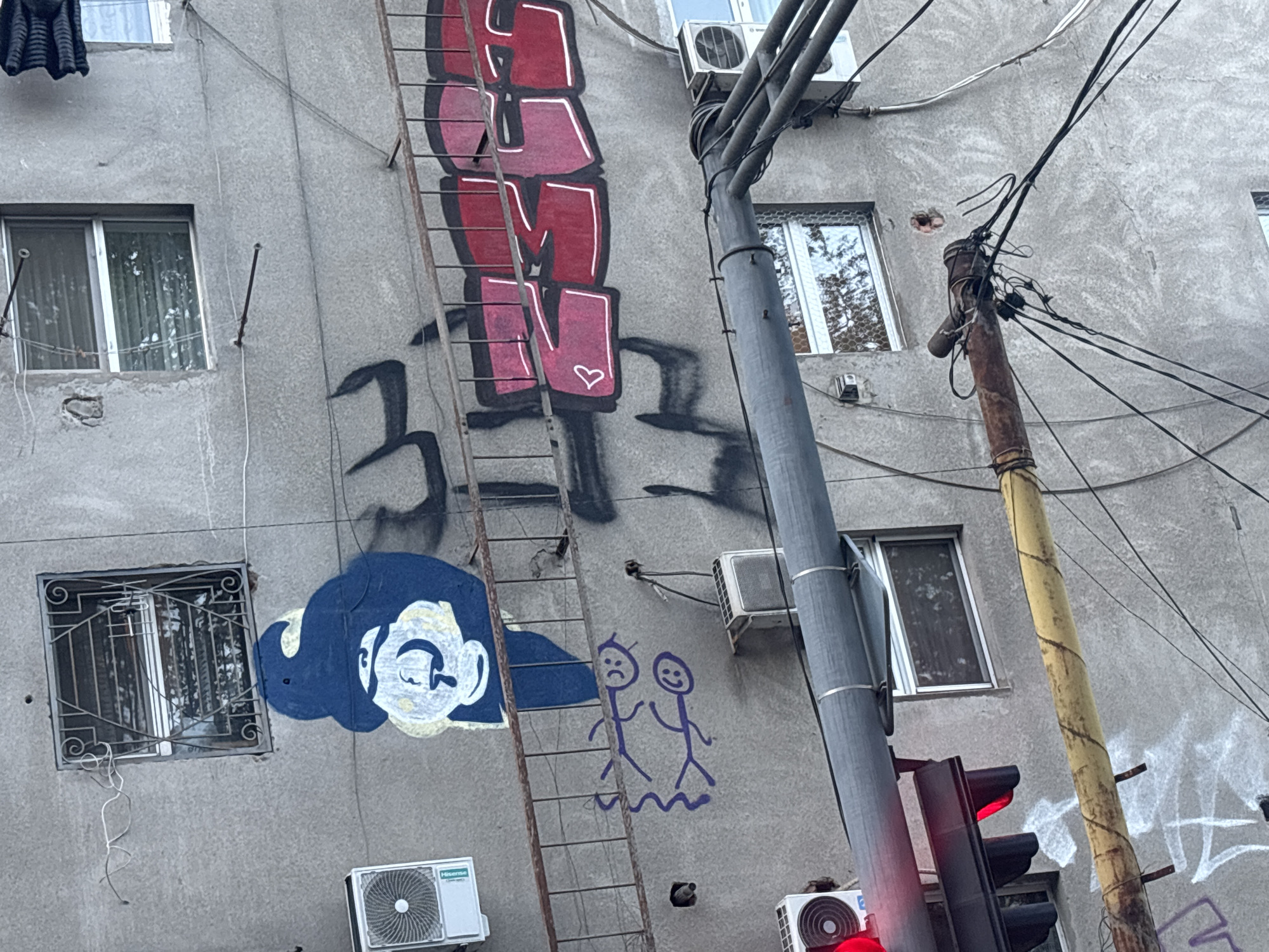 An outdoor, slightly low-angle photograph captures a weathered concrete building facade in Tbilisi, Georgia, characterized by urban graffiti and infrastructure. The multi-story building features several windows, some plain and others secured with metal bars. Dark laundry hangs from a line near an upper window.
Prominent graffiti includes a large red "CMN" with a white heart on the letter 'N'. Below this, a blue and white stylized head with closed eyes is depicted. To the right of the blue head, two purple stick figures are drawn, one with a sad expression and the other with a happy one, both standing on a wavy line. Other abstract black shapes and some indistinct white/purple graffiti are also visible on the grimy wall.
A rusty metal ladder is affixed vertically to the building. Numerous utility lines are strung across the scene, supported by a thick gray pole on the right, which partially obscures a red traffic light. Another rusty, yellow-wrapped pole with more wires stands further to the right. Several air conditioning units are mounted on the building, including a "Hisense" brand unit on the lower left. The lighting indicates it is daytime, possibly overcast.