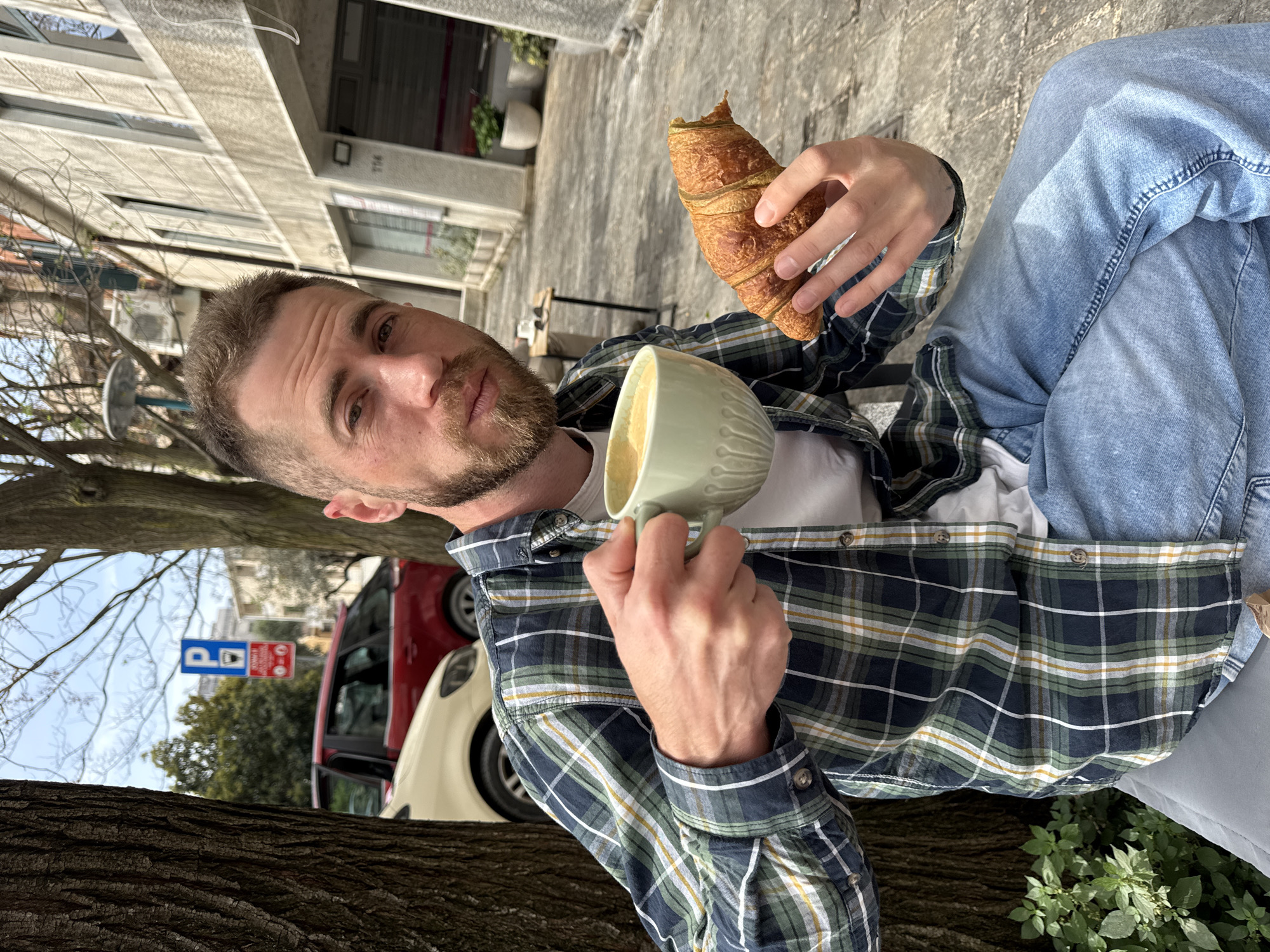 A man is seated outdoors, likely in a cafe or on a bench in Budva, Montenegro. He is holding a croissant in his right hand and a cup of coffee in his left. He is wearing a plaid shirt, a white t-shirt, and blue jeans. A parking sign is visible in the background, as well as a red and white car. The image is rotated.