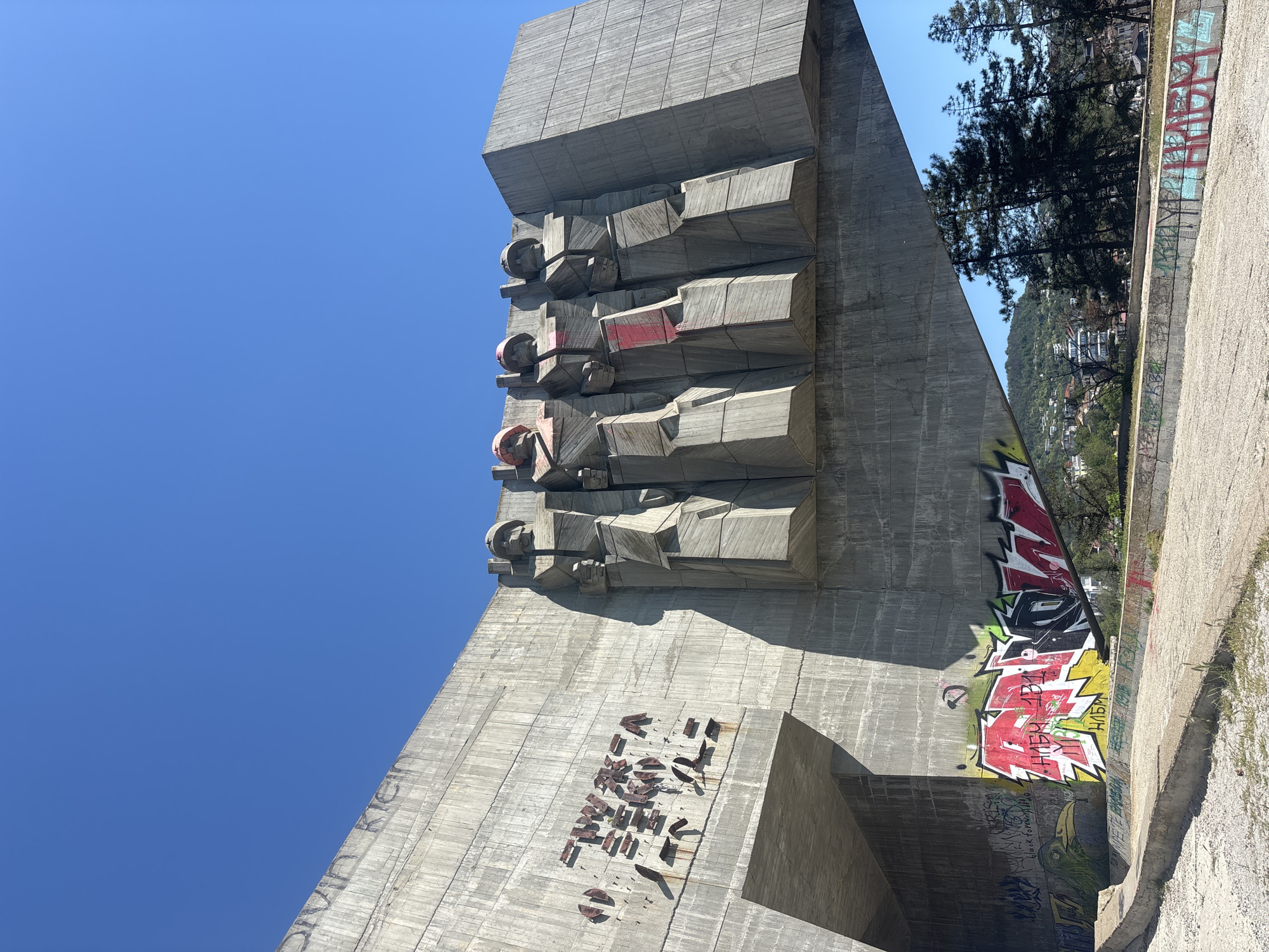 The image shows a tall, concrete monument in Varna, Bulgaria, set against a clear blue sky. The monument is angular with a series of cubic protrusions near the top, some partially painted red. The lower part of the structure has missing metallic ornamentation. Graffiti is visible on the right side of the monument and on the wall adjacent to it, consisting of red, black, yellow and green letters and shapes. Trees and a view of buildings on a hillside can be seen behind the wall to the right of the monument.