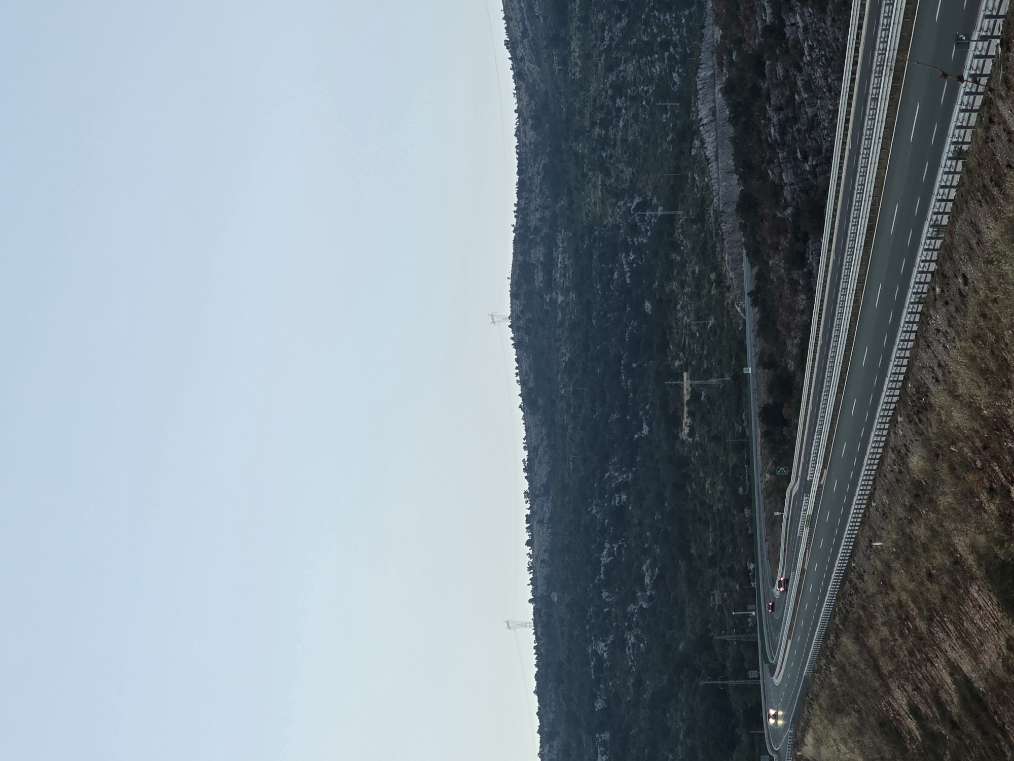 An elevated, wide-angle shot captures a modern highway curving through a rugged, sparsely vegetated landscape under a pale, overcast sky. The highway, with multiple lanes separated by barriers, is the central focus, with guardrails running along its edges. Two cars, with their headlights illuminated, are visible on the highway, suggesting it is dusk or dawn.
The terrain flanking the highway is characterized by dry, brown earth and sparse, low-lying vegetation in the foreground, transitioning to a dense, dark green forested hillside in the background. The hillside is dotted with rocky outcrops and appears to be a natural, undeveloped area. In the distance, atop the ridge of the hillside, several tall, lattice-like electricity pylons are visible against the sky, indicating the presence of power infrastructure. The overall scene is serene and devoid of human activity beyond the vehicles on the road. The location is identified as Klopot, Montenegro.