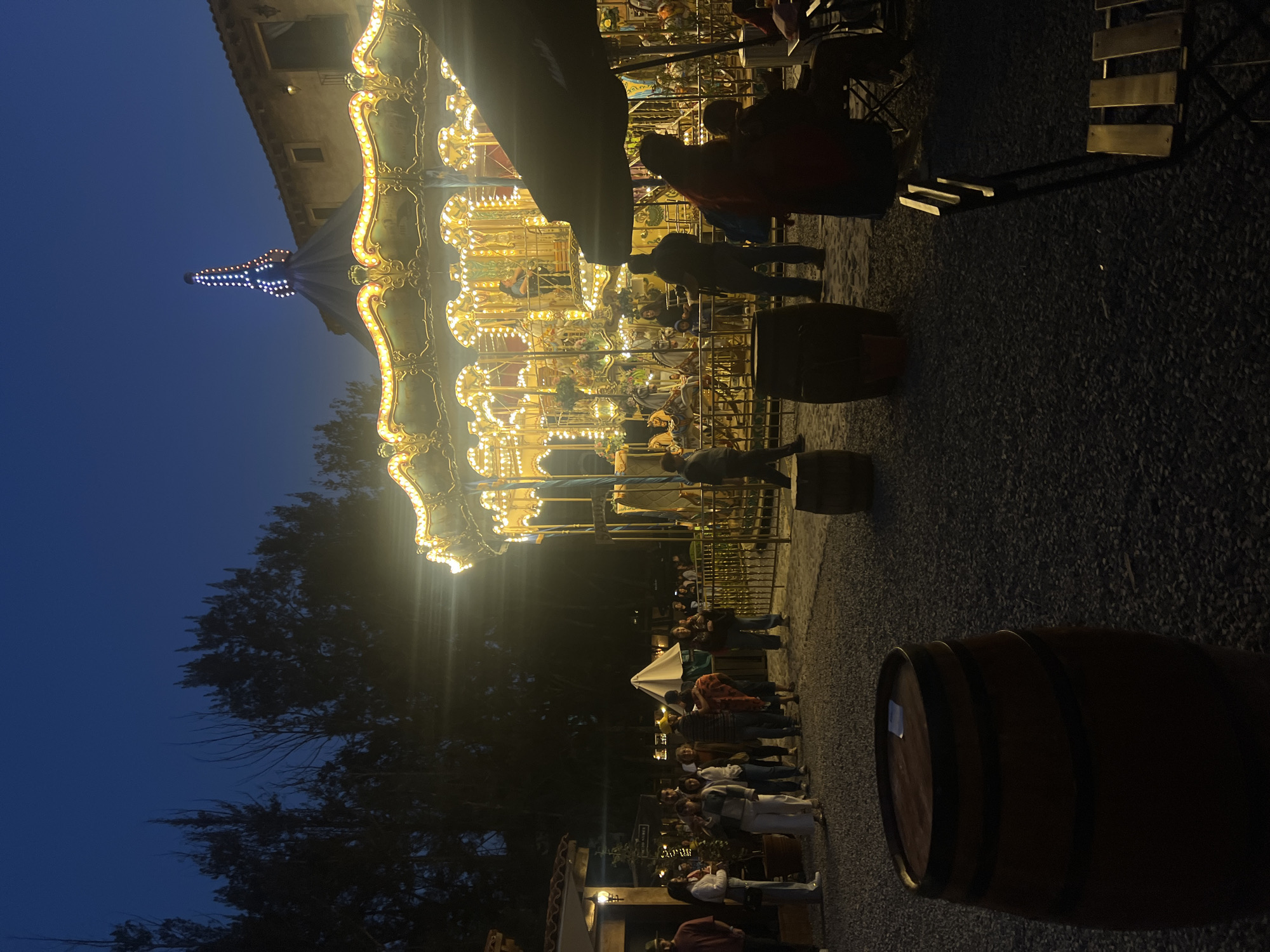 The image depicts a carousel illuminated at night in Domingo Arenas, Mexico. The carousel is brightly lit with numerous decorative lights. A crowd of people surrounds the base of the carousel, some standing and observing, while others appear to be near the ride. In the foreground, there are several wooden barrels. Chairs and tables are nearby the carousel, indicating a seating area. Trees are visible in the background, silhouetted against the dark blue night sky. The ground is covered in gravel.