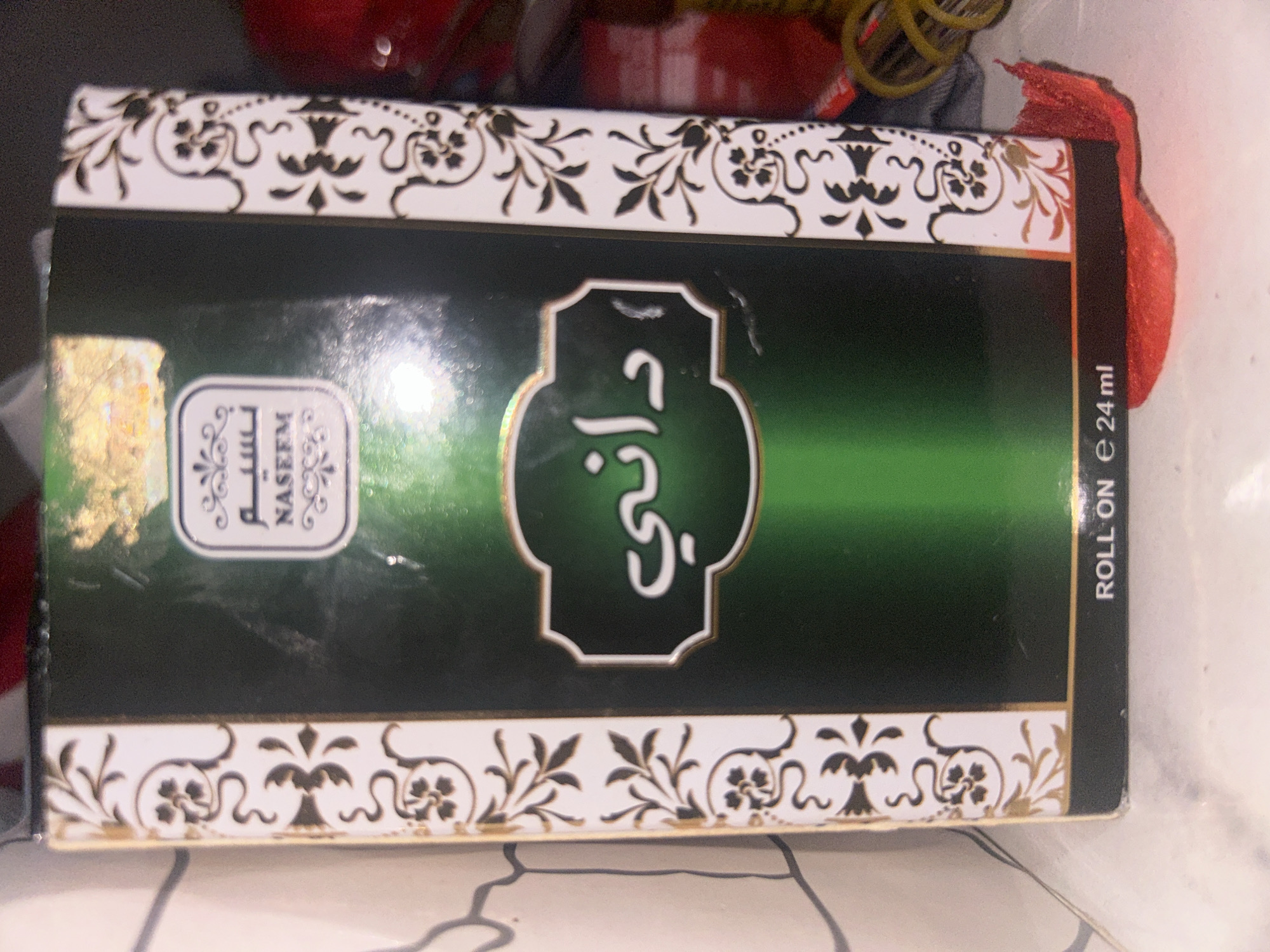 The image shows a close-up of a perfume box. The box is primarily dark green with ornate white and gold floral designs on the sides. In the center of the front panel, there is a white, decorative frame containing Arabic text and the word "NASEEM" in English. Below this, larger Arabic text is visible in white within a similar frame. At the bottom of the box, text reads "ROLL ON e 24 ml". The box appears to be placed on a surface that is either a marble countertop or a light-colored tiled surface. The lighting suggests an indoor environment. There is no indication of the city of Ogbomosho, Nigeria, or any people in the image.