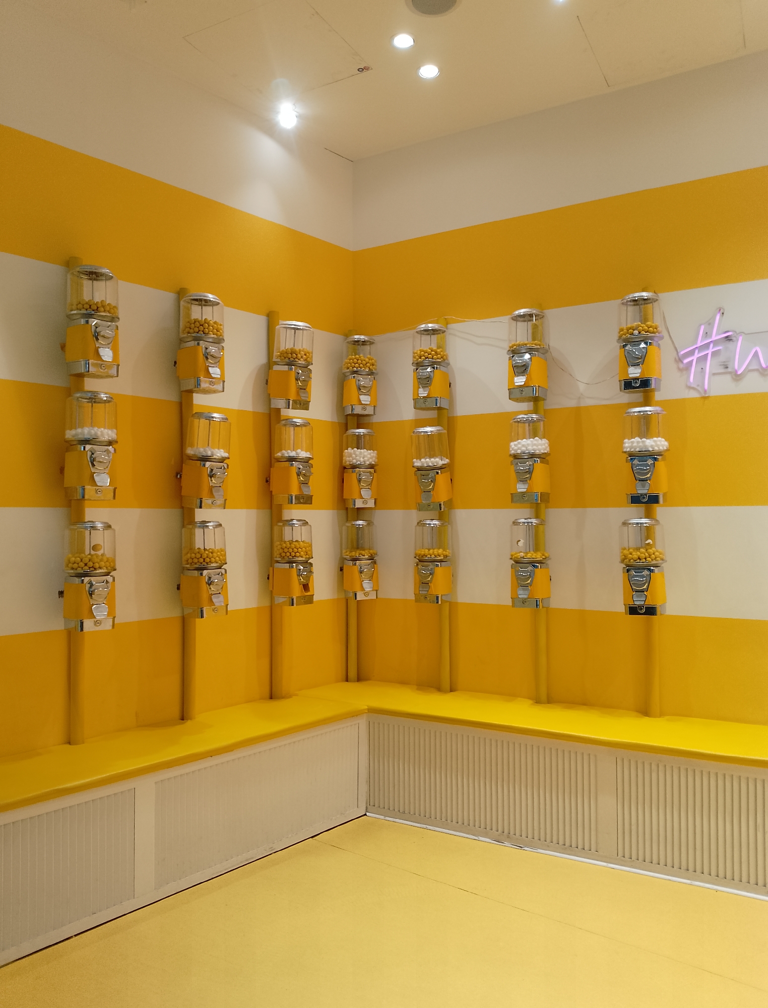 The image depicts a retail store interior with a vibrant yellow and white striped wall. Mounted on the wall are multiple yellow and chrome gumball machines, each filled with either yellow or white gumballs. The gumball machines are arranged in vertical columns, with four machines per column, and there are four columns visible. A neon pink sign with the hashtag symbol and the letters "w" is mounted on the wall to the right of the gumball machines. The floor is a bright yellow, and a yellow bench with a ribbed white base runs along the bottom of the wall, beneath the gumball machines. The scene appears to be well-lit, with overhead lighting visible in the ceiling. There are no people present in the image.