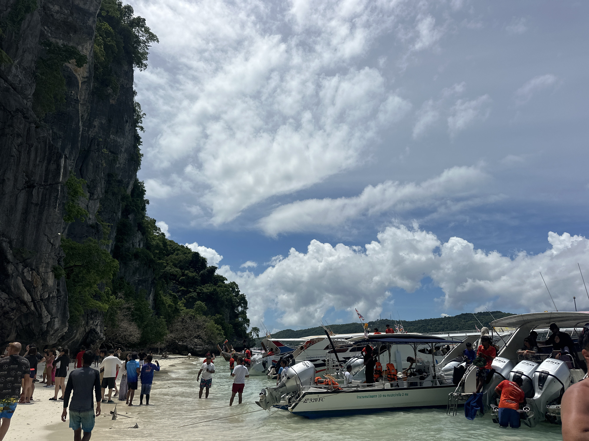 This media file captures a busy scene on a sandy beach in Ao Nang, Thailand. A tall, lush green cliff face dominates the left side of the frame, with the sea gently lapping at the shore. The water is clear and shallow, revealing the sandy bottom.
Numerous people are present, some wading in the water, while others are gathered on the beach. They appear to be a mix of tourists and locals, dressed in casual beachwear. The atmosphere suggests a typical tropical day, with a bright sky dotted with fluffy white clouds.
Several boats are moored in the shallow water, most of them are speedboats with outboard motors. Some of these boats are labeled with Thai text, one of which reads "จํานวนคนโดยสาร 10 คน คนประจำเรือ 2 คน", indicating passenger and crew limits. People can be seen boarding and disembarking these boats, suggesting that they are being used for tours or transportation. The presence of the boats and the activity around them point to a tourist destination, likely a popular spot for island hopping or excursions.
The time of day appears to be late morning or early afternoon, given the bright sunlight and the intensity of the shadows. The weather is warm and sunny, ideal for a day at the beach. The overall impression is one of a lively, active coastal environment, bustling with people enjoying the tropical beauty of Thailand.