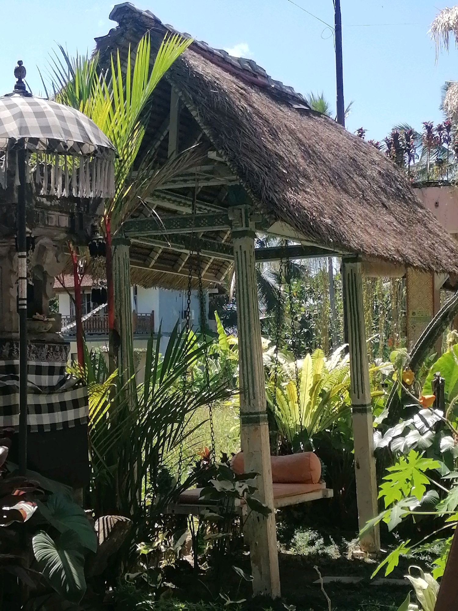 In the city of Ubud District, Indonesia, a thatched-roof pavilion stands amidst lush greenery under a clear blue sky. The pavilion, supported by four fluted columns, features an open-air design with a suspended swing adorned with an orange cushion. To the left of the pavilion, a checkered black and white umbrella stands beside a small stone altar. Dense tropical foliage, including palm fronds and ferns, surrounds the structure, creating a shaded and serene atmosphere. In the background, a light blue building with balconies is visible, hinting at a residential or hospitality setting. The scene captures a tranquil moment within a tropical landscape.