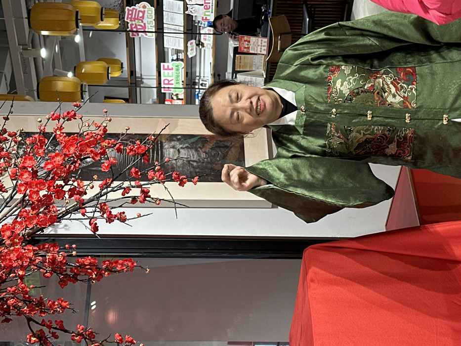 An adult East Asian female, estimated to be in her 50s to 60s, is centrally positioned, smiling broadly at the viewer in an indoor commercial or cultural establishment in Yuzhong District, China. She wears a dark green, traditional Chinese-style long-sleeved robe (hanfu or similar) featuring a white collar and intricate gold, red, and white brocaded patterns depicting mythical creatures and clouds on the chest, fastened with gold-colored frog closures. Her right arm is raised, with her hand forming a "mini heart" gesture by pinching her index finger and thumb together.
To her immediate left, a large object or structure draped entirely in bright red fabric occupies the lower-left foreground. Above this, and extending across the upper-left and center of the frame, is a dense arrangement of artificial red plum blossom branches. Behind the woman, a framed piece of dark artwork or mural is visible on a light-colored wall.
To the right of the woman and extending into the background, a commercial setting is visible. Multiple yellow, cylindrical lanterns hang from the ceiling. A wooden shelving unit stocked with various items and signs, including "FREE" and "宠爱 LET'S GO!" (Love LET'S GO!), is present. Further back, a male figure, likely middle-aged, is partially visible behind a counter, holding a phone to his ear. The overall environment suggests a well-lit, festive, or culturally themed interior.