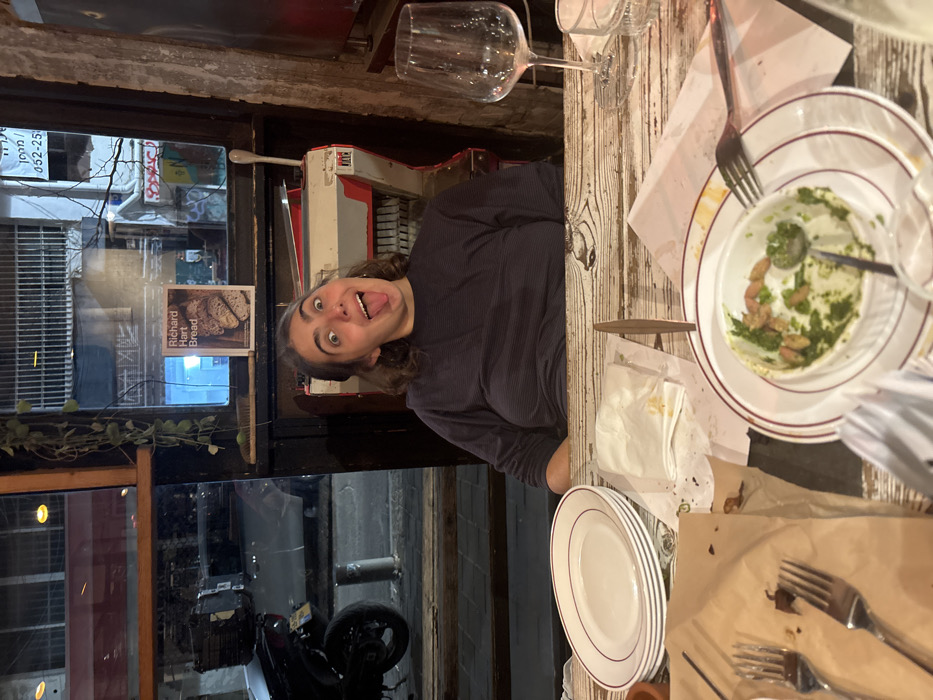 A person playfully sticks their tongue out next to a window displaying a "Richard Hart Bread" sign. The scene is at a restaurant, judging by the table setup with plates, forks, napkins, and wine glasses, some of which have remnants of food. The table appears worn. The photo's lighting is dim, possibly indicating an evening setting.