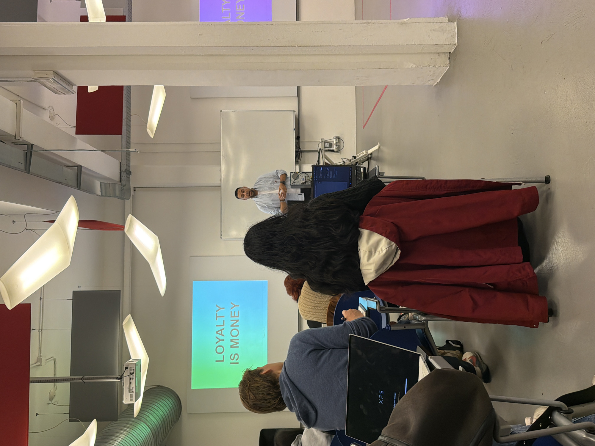 The image depicts an indoor presentation or lecture setting in what appears to be a modern, industrial-style space. A presenter stands in front of a whiteboard, facing a small audience seated in chairs.
Several attendees have laptops or tablets open in front of them. A projection screen displays the words "LOYALTY IS MONEY." There is exposed ductwork and modern lighting fixtures in the ceiling. The location is not identifiable as the city of Milan, Italy, solely from the visual information available in the image.