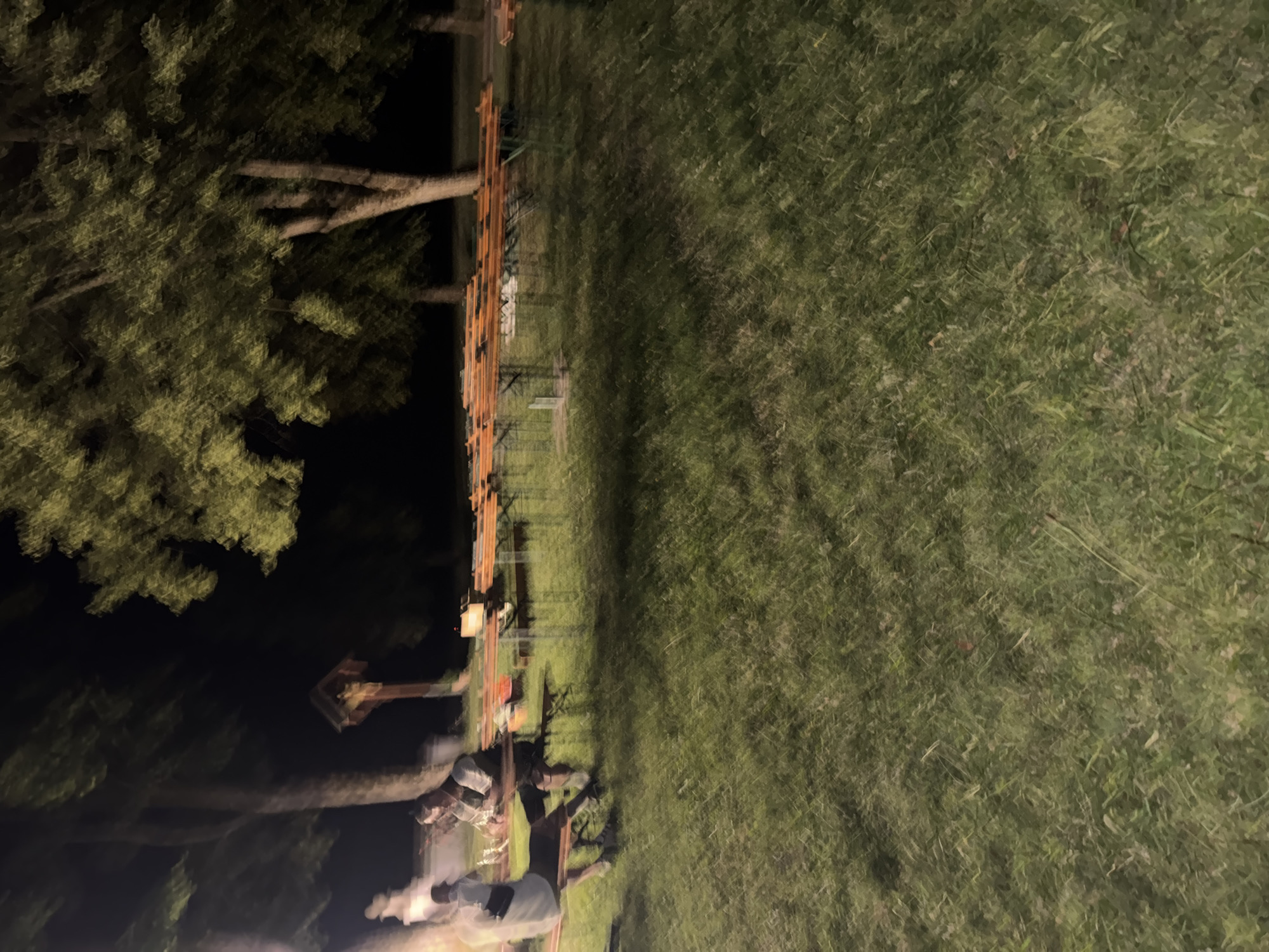 The image shows an outdoor gathering at night in Höhenkirchen-Siegertsbrunn, Germany. Several rows of wooden picnic tables and benches are set up on a grassy area. In the foreground, two people are seated at a table, with one facing away from the camera and the other looking towards the camera. A small wooden cross with a figure on it is visible in the background. The overall scene suggests a social event or a gathering at a park or beer garden. The lighting is dim, indicating it is nighttime, and there is a visible light source on the left side of the frame.