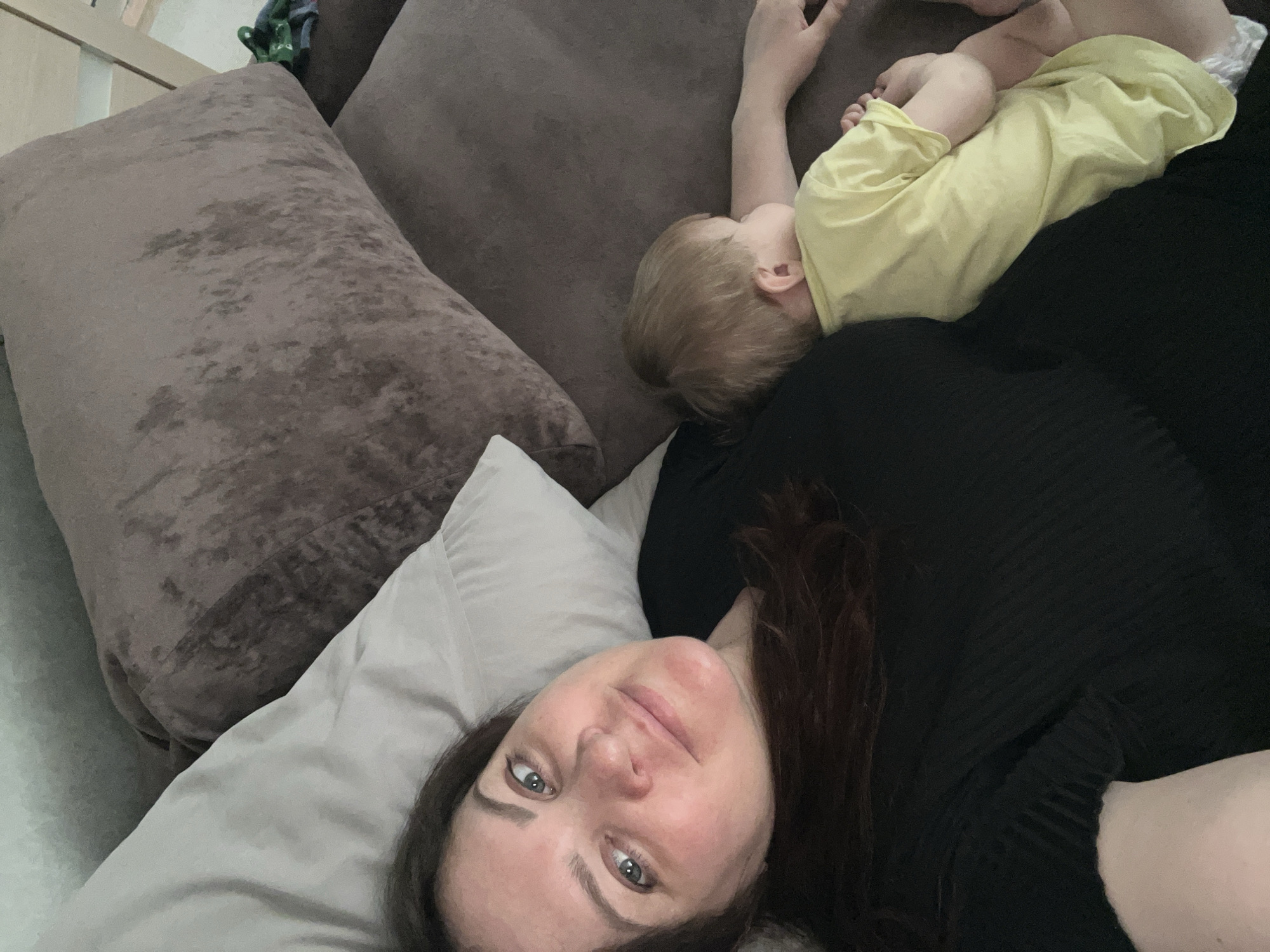 A woman with dark hair is lying on a gray pillow on a couch. She is wearing a black shirt and has gray eyes. A child wearing a yellow shirt is lying on the couch next to her. The couch has brown cushions. The location is likely inside a home. There is insufficient information to determine if the location is Noviy, Russia.