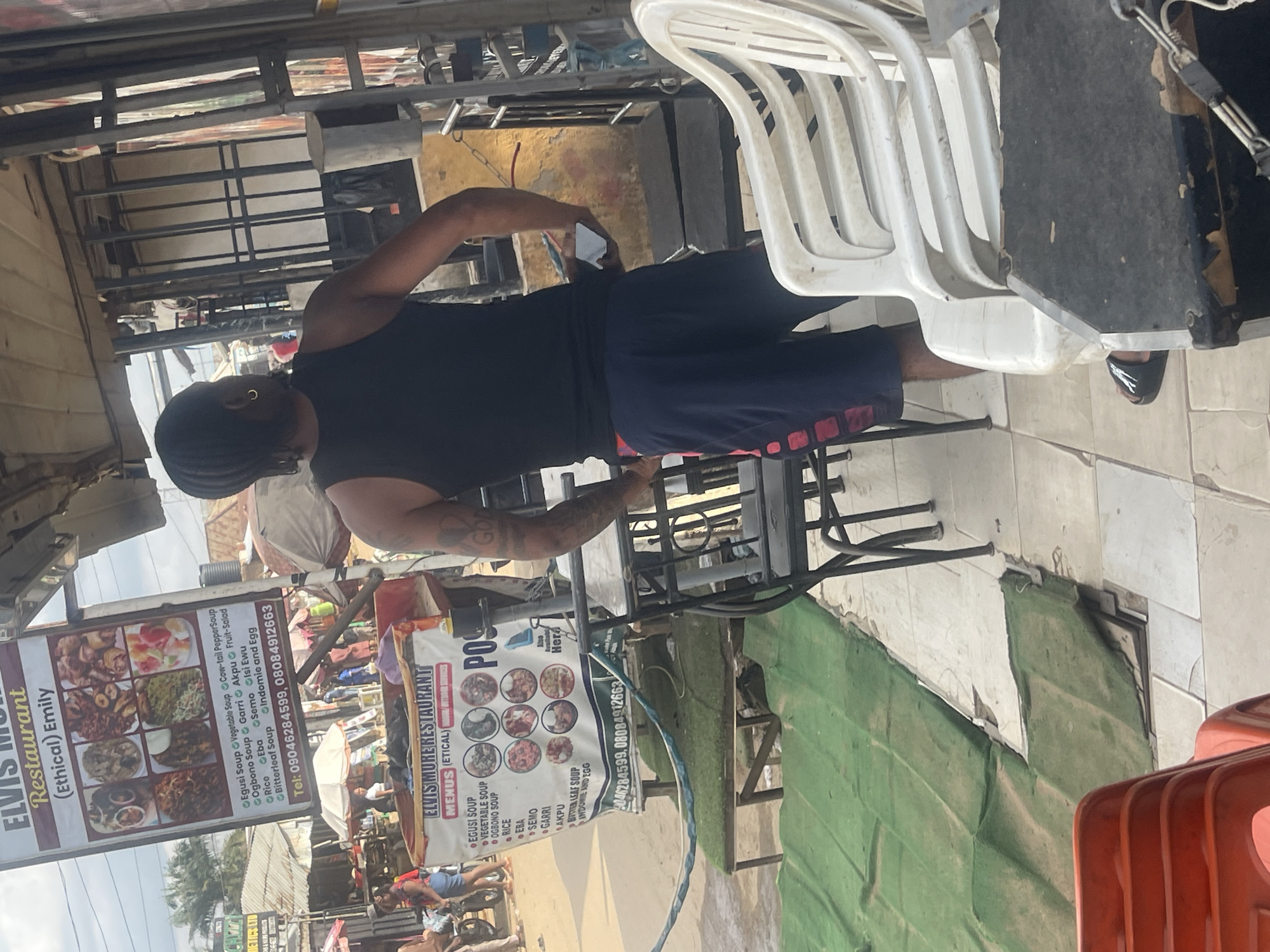 An outdoor view of a busy street scene in Abuja, Nigeria, features a man in a black tank top and navy shorts standing with his back to the camera, holding a white object. To his left, a restaurant sign for "ELVIS MORE RESTAURANT MENUS (ETICAL)" displays pictures of various dishes and lists menu items like "Egusi Soup," "Vegetable Soup," and "Rice." In the foreground, red and white plastic chairs are visible, some stacked. A green artificial turf covers a portion of the ground. In the background, other stalls and people can be seen, suggesting a market or commercial area. The overall impression is of a lively, urban environment.