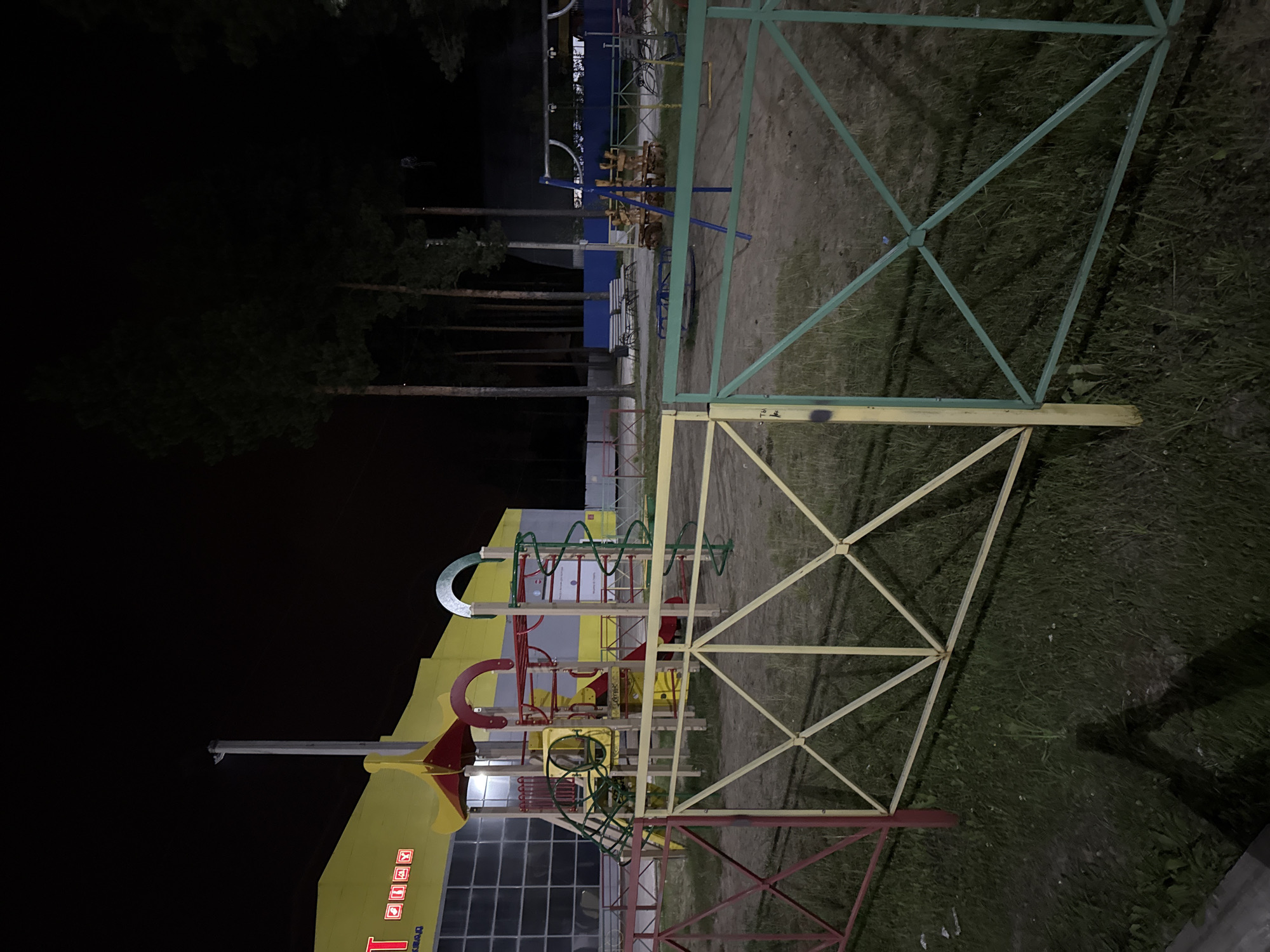 This is a nighttime, eye-level, medium shot of an outdoor children's playground located in Sovetskiy, Russia. The playground features various structures, including a slide, swings, and a merry-go-round. A large, yellow building with a dark, reflective facade stands behind the playground. Pine trees are visible in the background, and a fence with red, yellow, and green sections surrounds the playground. The ground is a mixture of sand and grass. The scene is illuminated by artificial lighting, casting long shadows. No people are present.