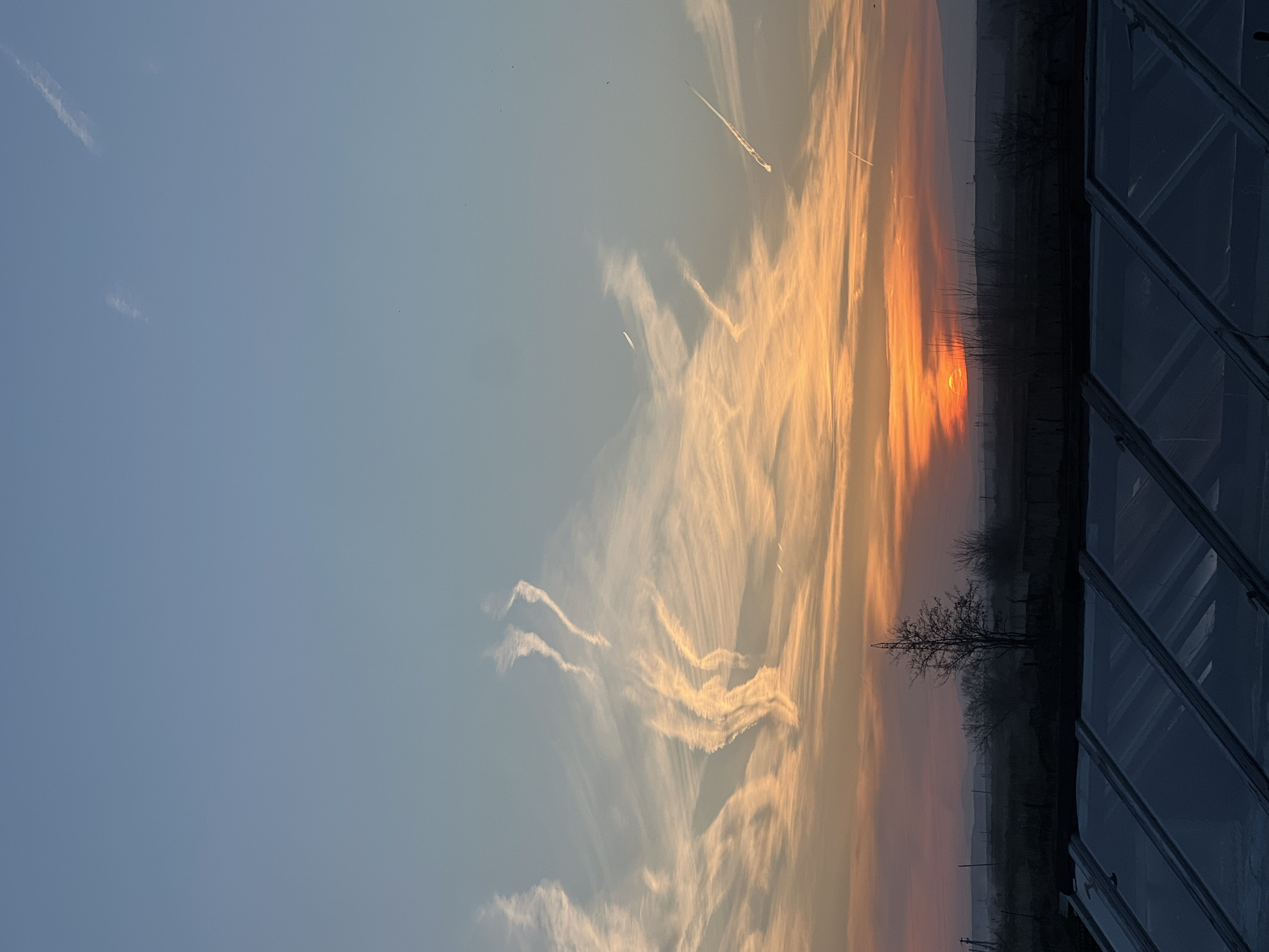The image captures a sunset scene in Azatan, Armenia. The sky is a gradient, transitioning from blue to orange as the sun sets on the horizon. Streaks of clouds are visible, reflecting the colors of the sunset. Silhouetted against the fading light are trees and vegetation, suggesting a rural or semi-rural setting. A structure, possibly a building or greenhouse, is visible on the right side of the frame. The general atmosphere is serene and tranquil.