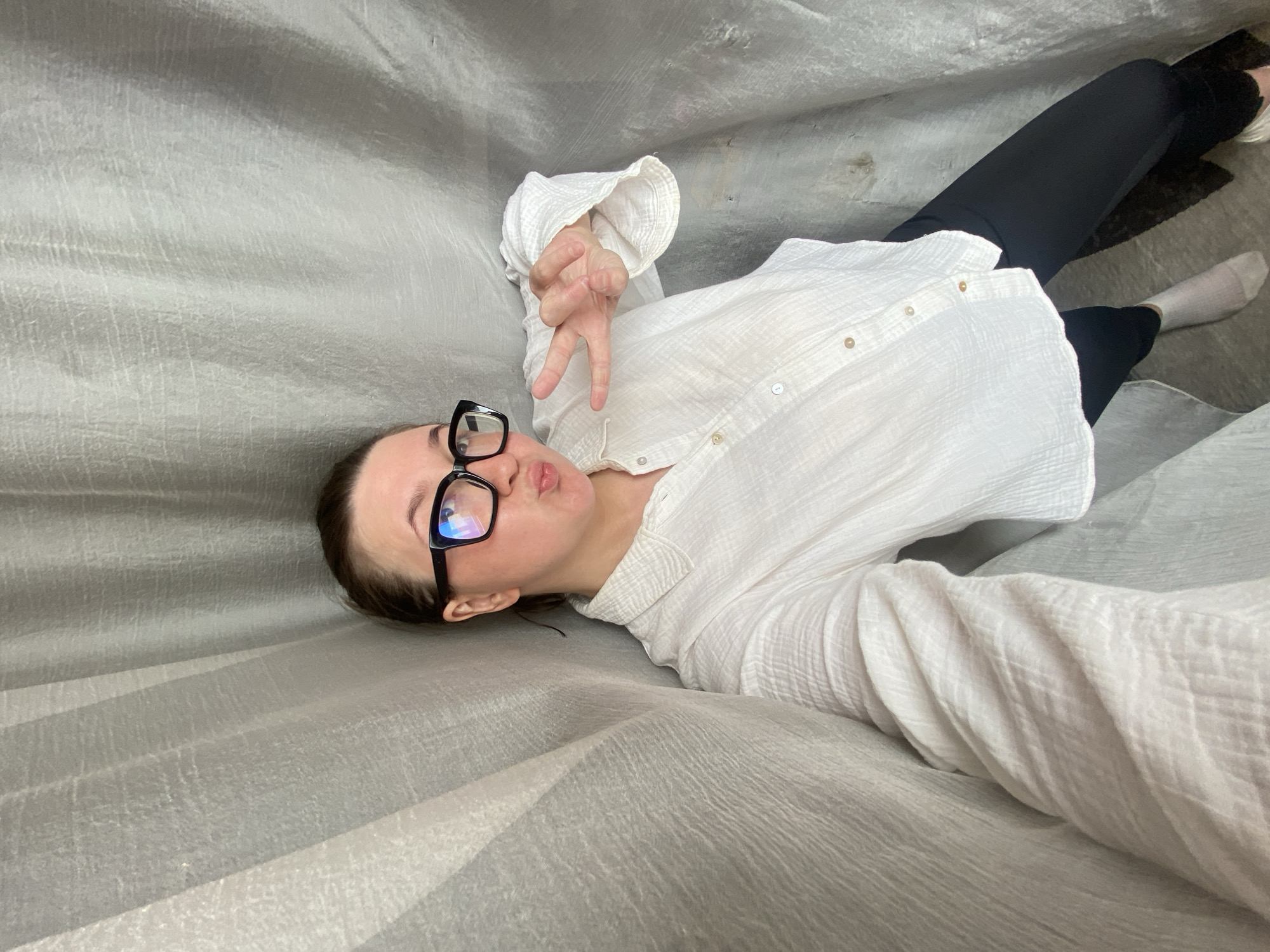 A young adult female is captured in a selfie, lying down or reclining against a heavily textured, wrinkled grey fabric surface, likely indoors in Chaikovsky, Russia. She is positioned diagonally across the frame, looking towards the camera with her head tilted to the right. She wears a white, button-down, long-sleeved shirt with a crinkled or waffle-knit texture, black trousers, and white socks. Black-framed glasses are perched on her face, reflecting light. Her lips are slightly puckered, and she extends her right hand towards the camera, forming a peace sign with her index and middle fingers. The background consists entirely of the grey, unevenly draped fabric, which shows subtle variations in light and shadow, indicating folds and creases. There is no visible text in the image. The overall impression is casual and playful.