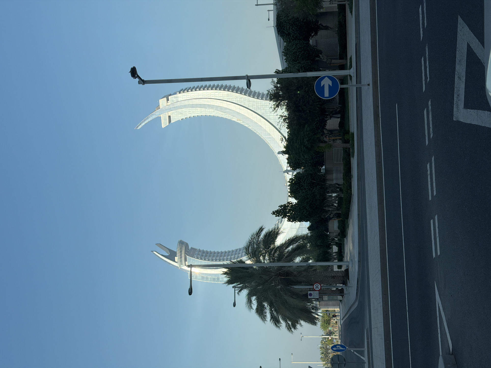 A wide shot of the iconic crescent-shaped buildings in Doha, Qatar, against a clear blue sky. In the foreground, a dark asphalt road with white lane markings dominates the lower portion of the frame. A sidewalk with grey paving stones runs along the right side of the road. Beyond the sidewalk, several palm trees and manicured trees are visible. Two prominent streetlights are positioned along the road. Traffic signs are present, including a circular sign with the number "30" inside a red border, and a blue circular sign with a white right-pointing arrow. The two distinctive modern buildings, with their white, curved facades and glass panels, rise dramatically in the background, creating a dramatic architectural statement. The overall scene is clean and modern, suggesting an urban environment.