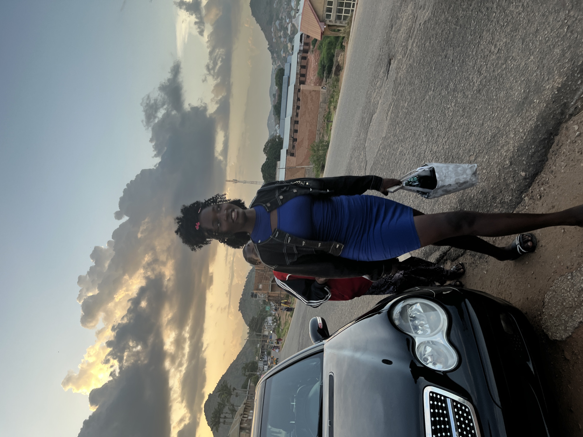 In Jos, Nigeria, on a dusty, paved road, a young woman with dark, curly hair adorned with a pink flower walks alongside a black Mercedes-Benz. She is wearing a form-fitting, bright blue dress with a cutout at the chest and a black leather jacket. She carries a light-colored patterned bag in her right hand. Behind her, partially obscured by her presence, another person wearing a red and black jacket and dark pants walks in the same direction. The background reveals a cityscape with buildings and hills under a dramatic sky filled with dark, illuminated clouds and hints of sunset colors. Palm trees and scattered buildings are visible on the slopes of the hills. Traffic, including cars and what appears to be a motorcycle, can be seen on the road in the distance. The overall scene suggests an outdoor environment on the outskirts of the city as evening approaches.