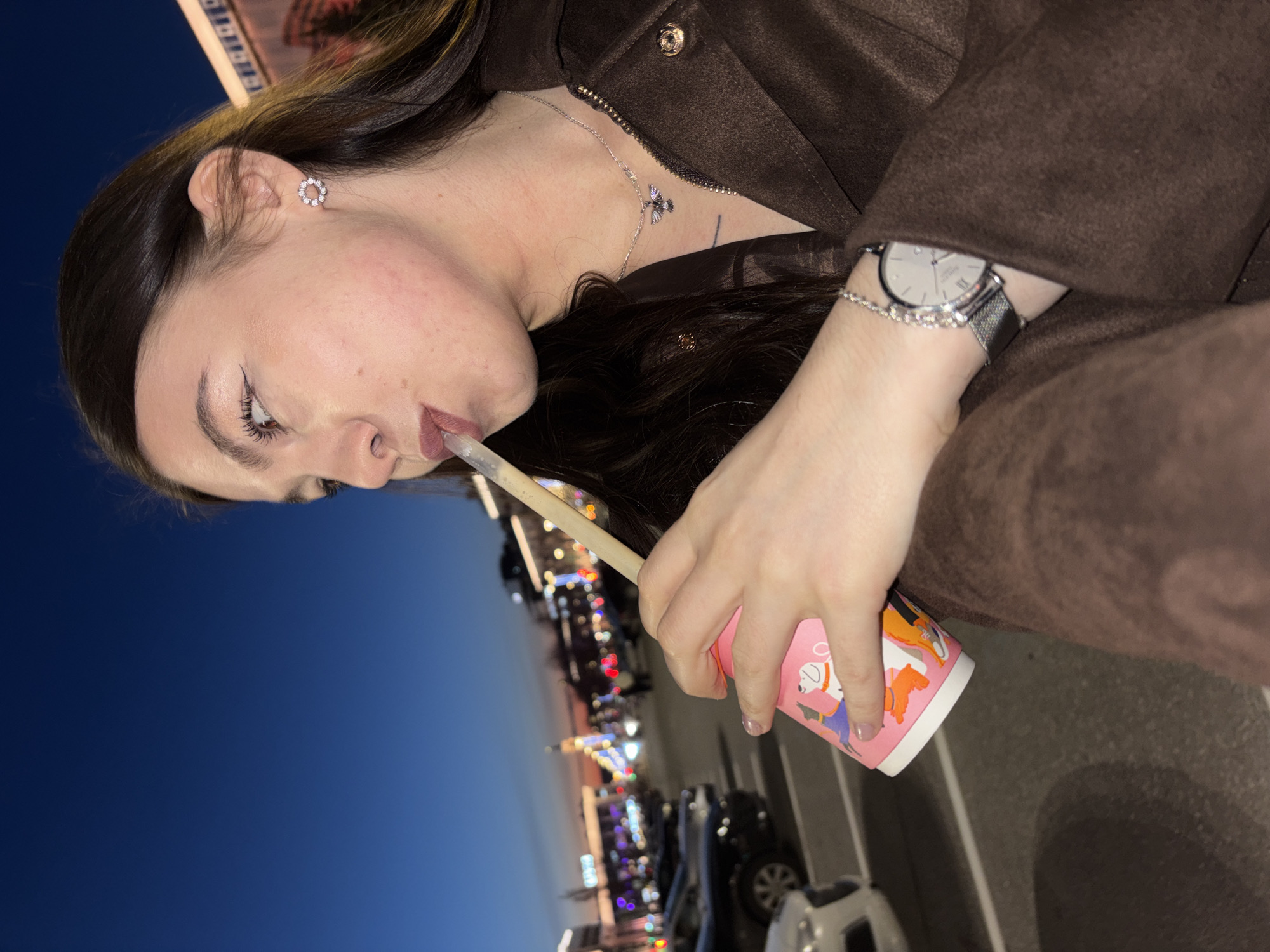 A woman is drinking from a pink cup with illustrations of dogs through a straw. She has a silver watch on her left wrist, a silver necklace, and earrings. She is wearing a brown jacket. In the background is a city street at dusk with illuminated buildings and parked cars. The location is identified as the city of Perm, Russia.