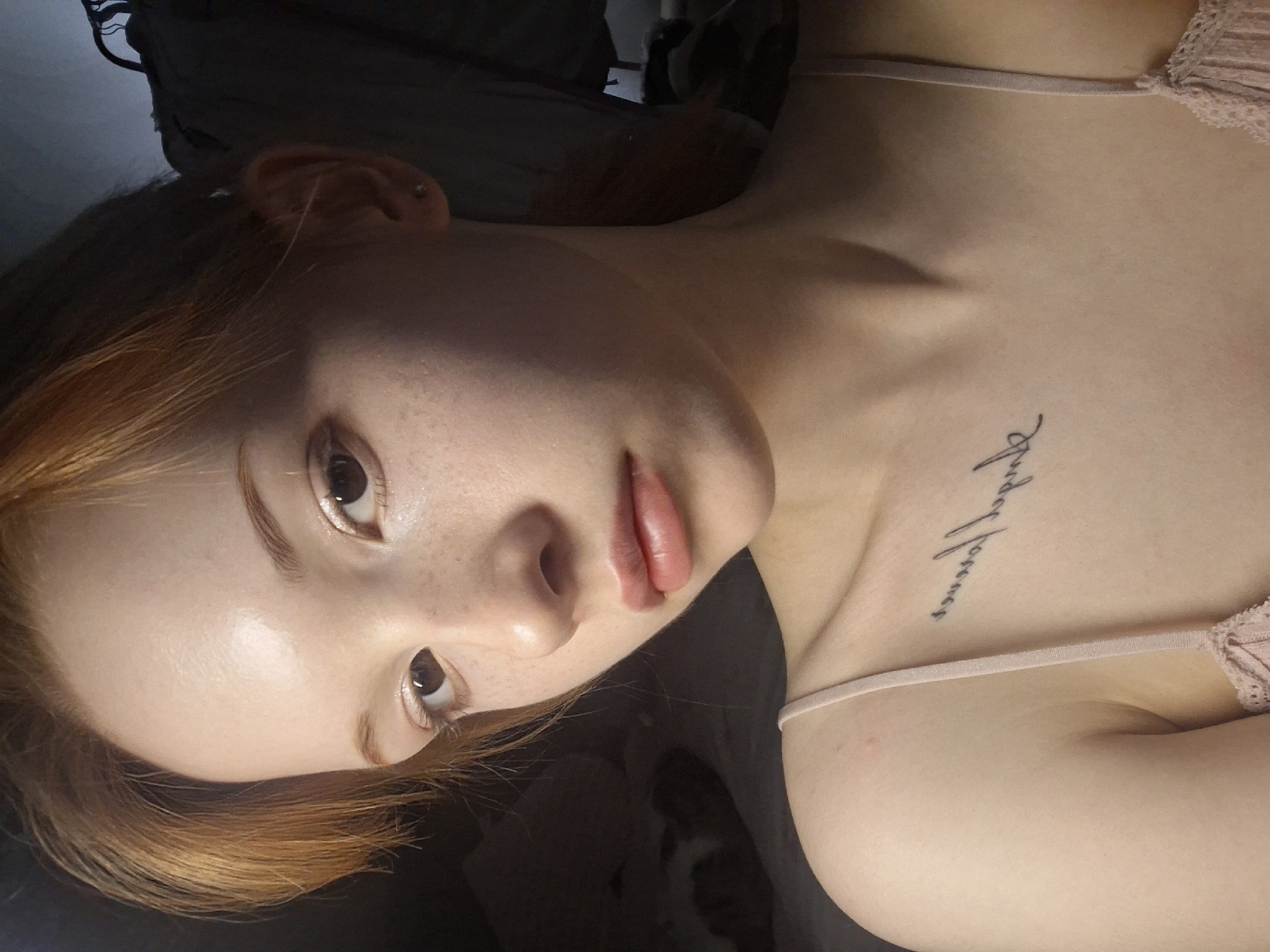 A young woman with red hair and freckles is lying down. She has a tattoo on her upper chest. She is wearing a light pink top with lace. The background is dark and indistinct. The photo does not contain enough information to confirm location in Chaikovsky, Russia.