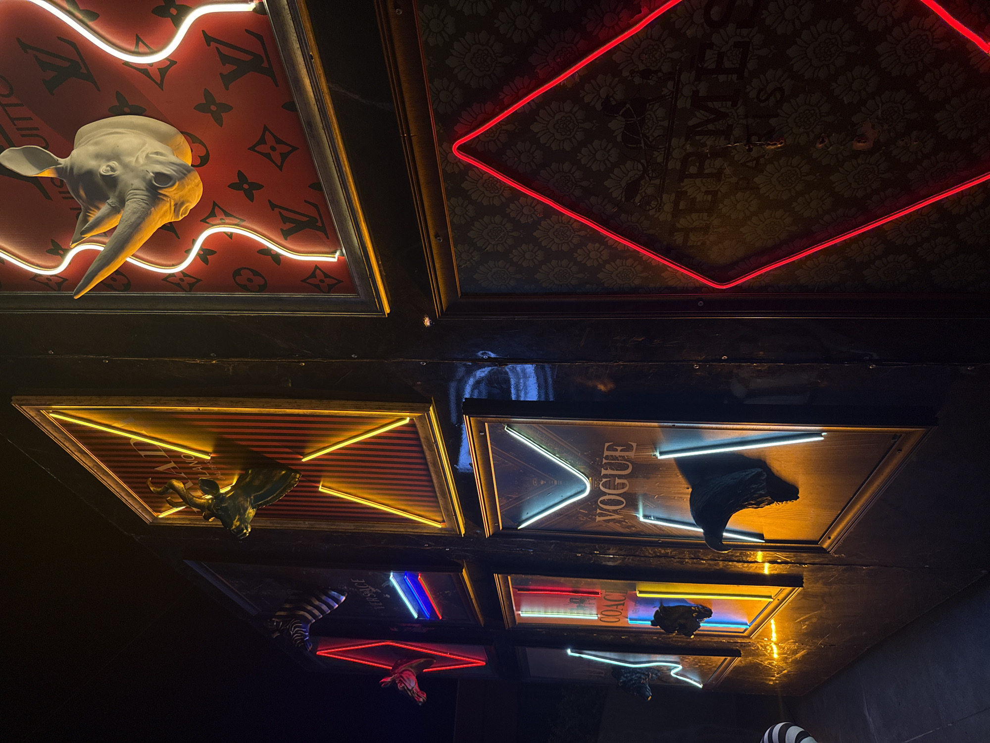 The image depicts a mirrored ceiling featuring several framed designs in Kemer, Turkey. The designs consist of a Louis Vuitton monogram background with a protruding white rhinoceros head and a white neon squiggle, a red diamond-shaped neon light surrounding a floral pattern, a striped background with a gold-colored bull head and gold neon lines, a "Vogue" logo with white neon lines and a black rhinoceros head, and logos for "Versace" and "Coach" with corresponding neon lines and animal heads. The reflection shows other logos with corresponding lights and animal heads.