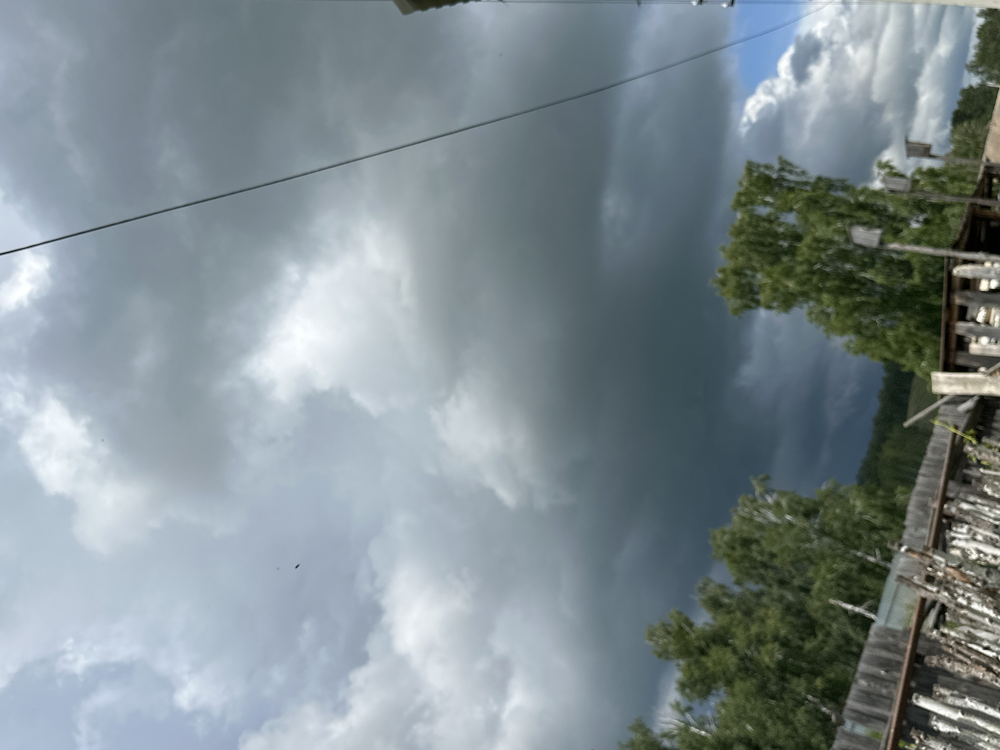 The image depicts a cloudy sky over a rural setting. The sky is dominated by heavy, dark storm clouds, with some lighter, fluffy clouds visible on the right side, hinting at a break in the weather. In the foreground, a wooden fence made of rough-hewn logs runs horizontally across the lower part of the frame. Behind the fence, there are trees with lush green foliage. A single power line diagonally crosses the sky from the upper right to the upper left corner. The overall scene suggests an overcast day, possibly before or after a storm. There are no people or animals visible. The location is described as Unknown, Unknown.