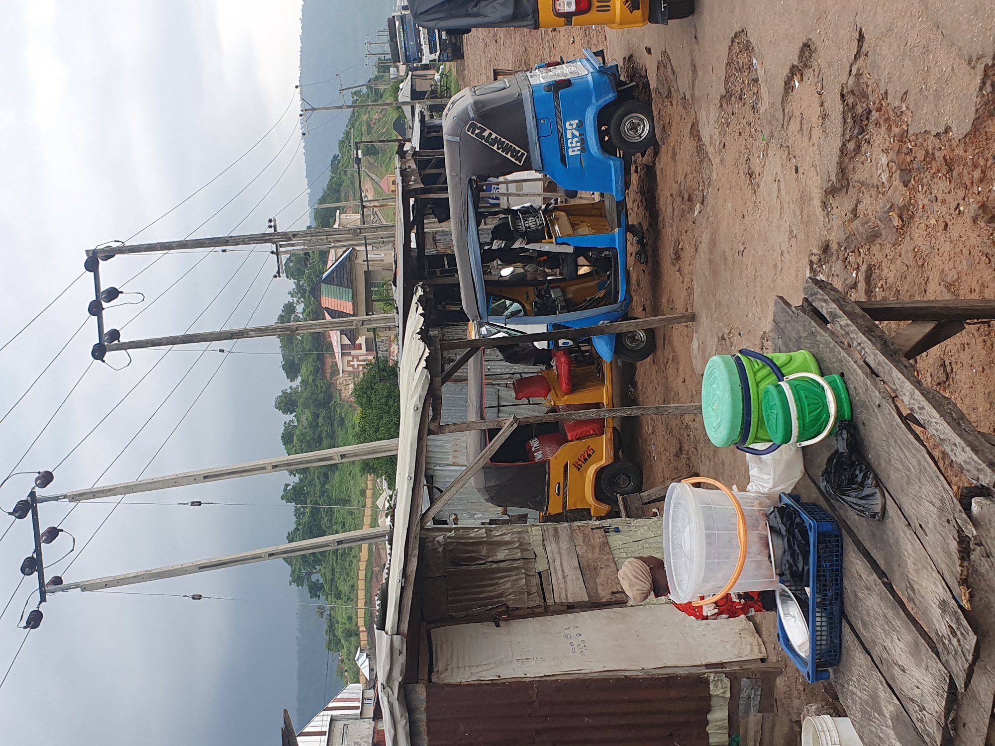 The image depicts a street scene in Lokoja, Nigeria, characterized by a roadside market and transportation hub. In the foreground, a rustic wooden table is laden with various items, including a blue plastic basket containing a metal plate and black plastic bags, a large clear plastic bucket with an orange handle, and two green buckets with lids. A person wearing a head covering and a patterned red and white garment stands behind the table, partially visible.
Behind the table, several tricycles, commonly known as "Keke Napep," are parked. One yellow tricycle, labeled "UNIMAS," is positioned to the left, while a blue and black tricycle with the license plate "R679" is prominently displayed in the center. To the right, the front of another yellow tricycle is visible. The structures surrounding the market are makeshift, constructed from corrugated iron and wood, suggesting a temporary or informal setup.
In the background, electrical poles with numerous wires stretch across the frame, indicating the presence of a power grid. Beyond the market, a hilly landscape covered in lush greenery is visible, with some buildings scattered on the slopes. The sky is overcast, contributing to a diffused lighting condition. The ground is a mix of dirt and gravel, with some areas showing signs of wear and tear, suggesting a less developed urban environment. The overall impression is one of a bustling local community engaged in commerce and daily transportation.