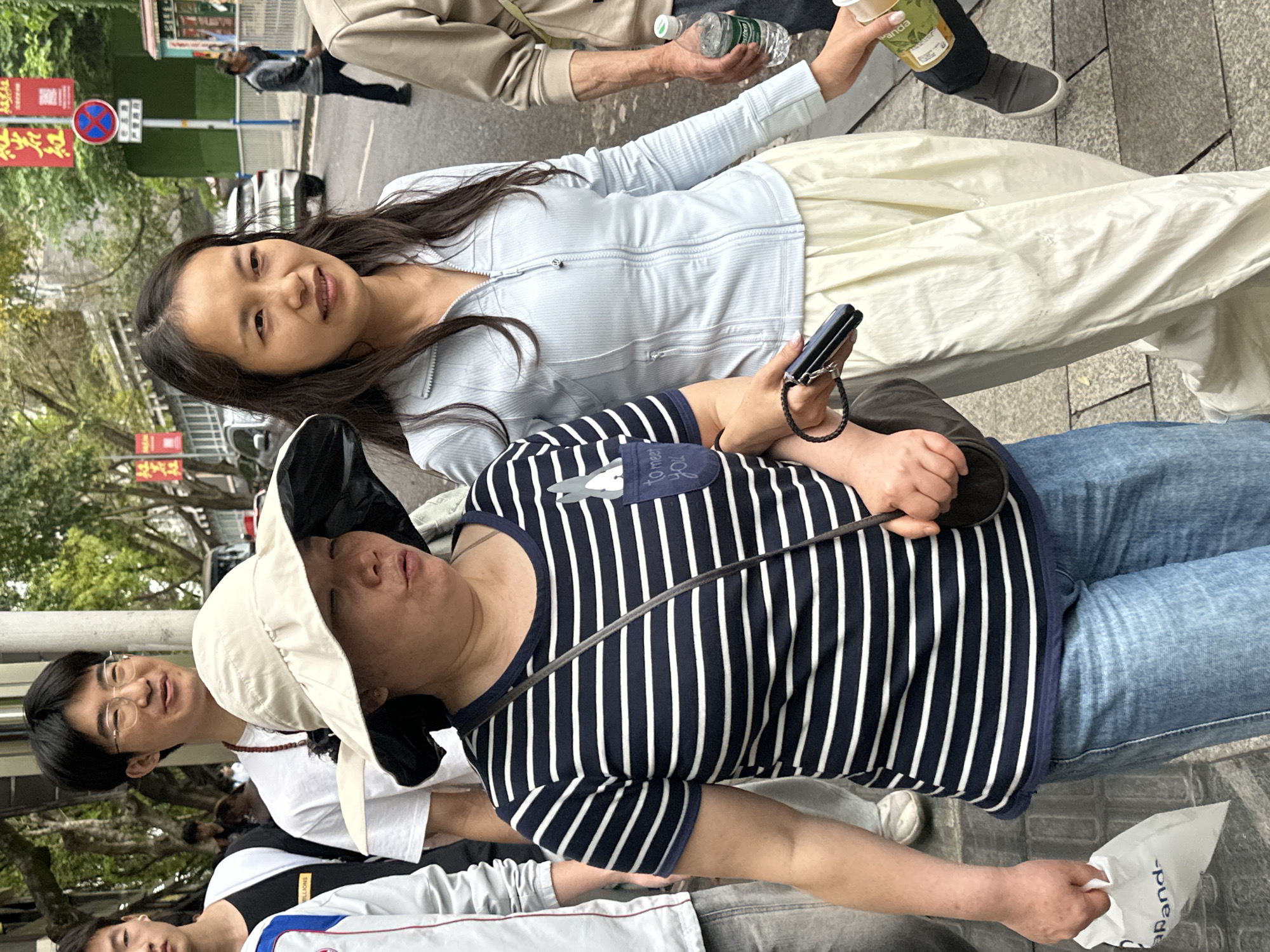 A street scene in Shapingba District, China shows a group of people. A woman with long dark hair wearing a light blue zip-up jacket and light-colored pants is smiling and looking at the camera. In front of her, a person wearing a navy and white striped shirt, light blue jeans, and a tan sun hat is holding a black cell phone in their right hand. The person is positioned at an angle, appearing to lean towards the right. Behind this person, another person wearing glasses and a white shirt with a beaded necklace is visible. Additional individuals are partially visible at the bottom and top of the frame. Buildings, trees, and street signs are in the background.