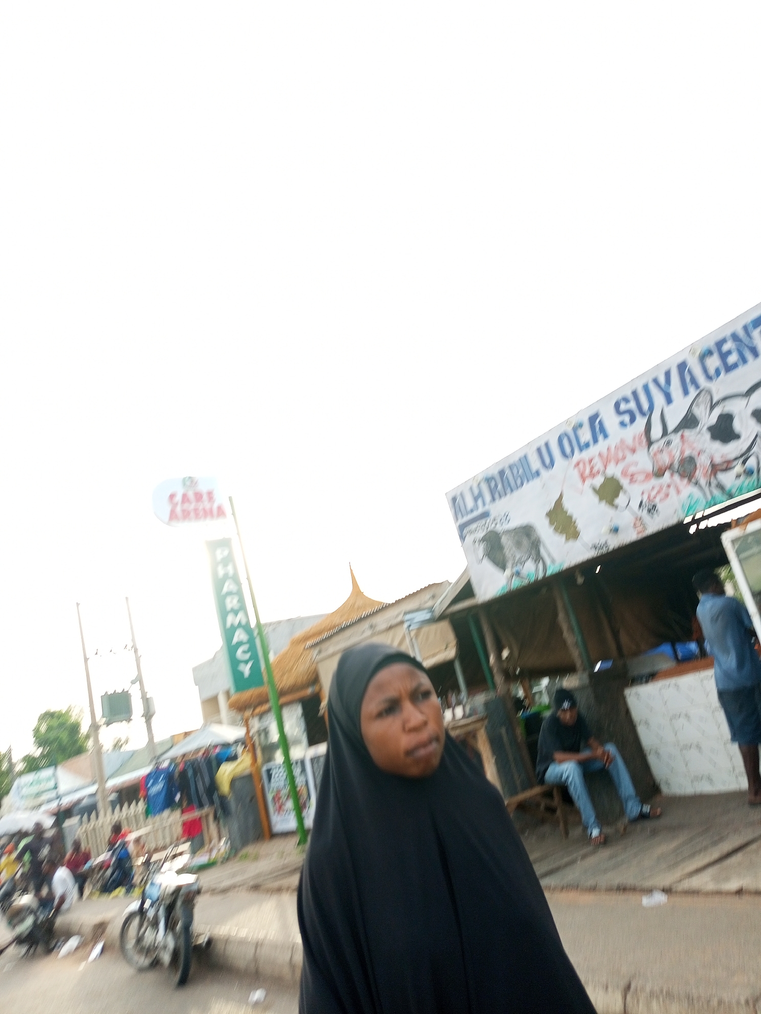 This is a street scene in Tempure, Nigeria, under a bright, overcast sky. A woman in a black hijab is in the foreground, looking to the left with a neutral expression. Behind her, a man in a blue shirt and shorts stands near a stall. To the right, another man in a black t-shirt and blue jeans sits on a wooden bench. In the background, a street market is visible with various stalls, including one with a sign that reads "ALH. RABILU OCA SUYA CENT." Another sign, partially obscured, indicates "PHARMACY." Several motorcycles and people are present at the market. The environment suggests a busy commercial area.