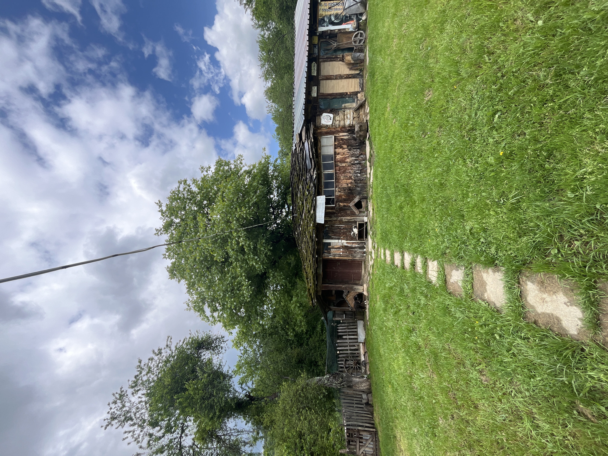 The image depicts a rustic, weathered building, likely a barn or storage structure, in Shopite, Bulgaria. The structure is constructed of wood, with a corrugated metal roof. It has a single visible window and a dark, weathered wooden door. The building is surrounded by lush green grass and trees, with a stone path leading towards the door. The sky is partly cloudy. A utility wire stretches diagonally across the upper left portion of the image. A wooden fence and various other farm implements can be seen partially obscured by vegetation.