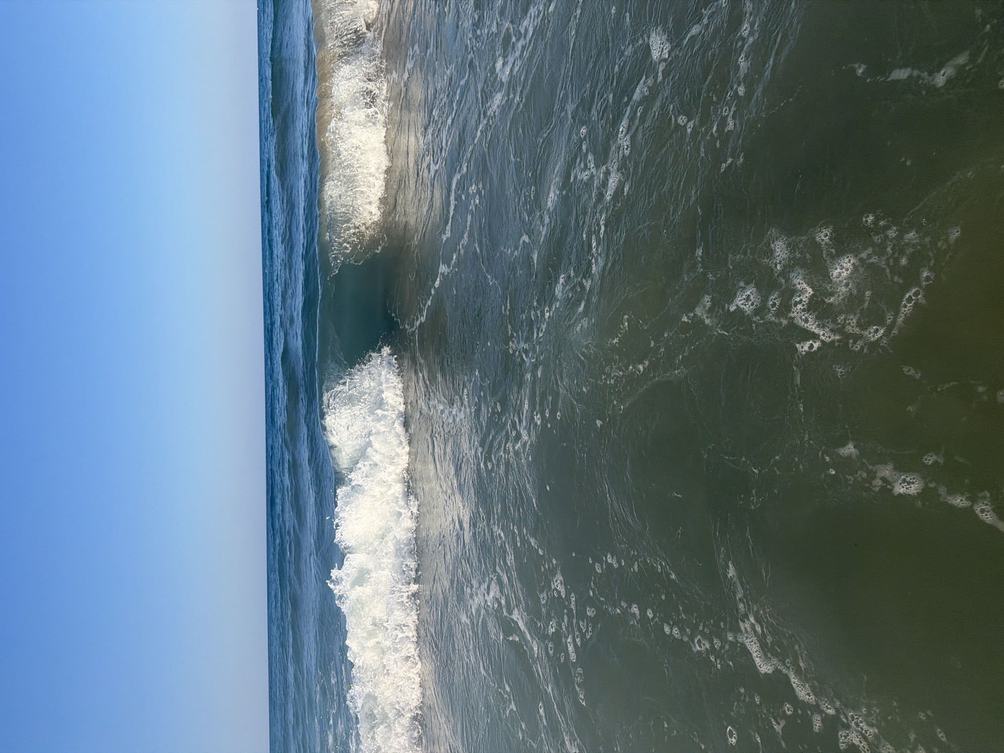 The image shows a close-up, aerial view of the sea and the sky. The scene is divided vertically, with a clear blue sky on the left and the ocean on the right. Two waves are visible in the ocean. The first wave is larger, breaking and creating white foam. The second wave, further out, is smaller and more defined. The water is a deep, dark green with lighter streaks and patches of white foam scattered across its surface. The overall impression is of a bright, clear day at the seaside. The image is oriented vertically, making the sky appear to the left of the water. No people, objects, or text are visible in the image.