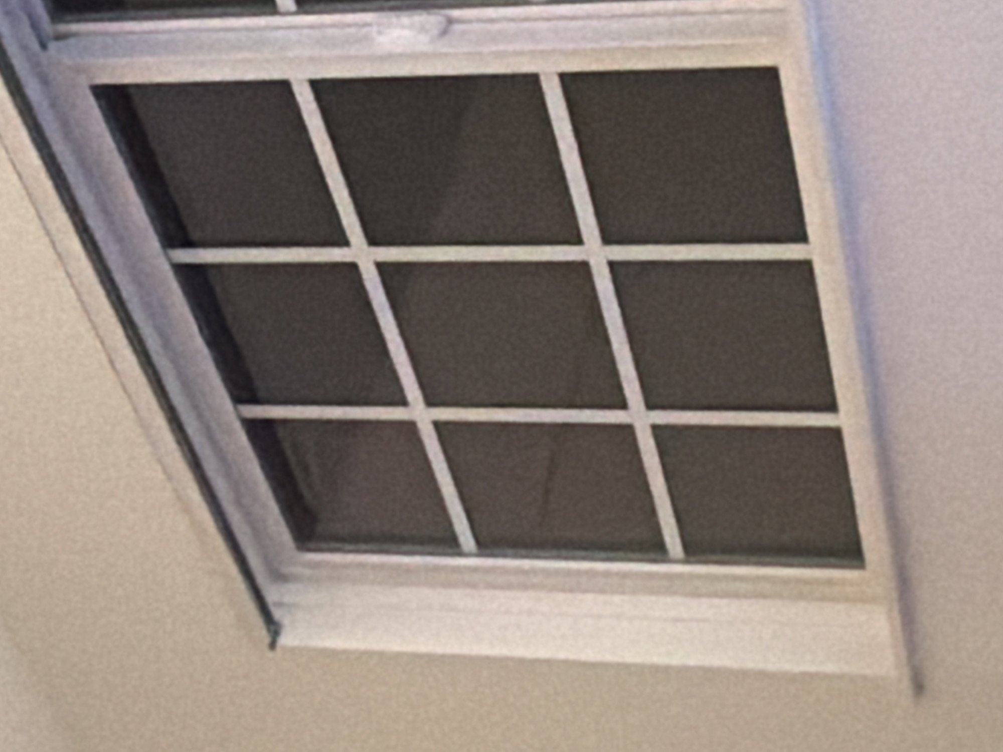 The image shows a portion of a window with a grid pattern. The window is framed by white trim and set against a light-colored wall. The glass of the window is dark, obscuring any view of the outside. The scene is indoors and appears to be a residential setting. There are no people or animals visible in the image, and no identifiable objects that would suggest a specific location in Portland, United States of America. The image is a close-up, focused on the window itself, with no contextual clues about the surrounding environment.