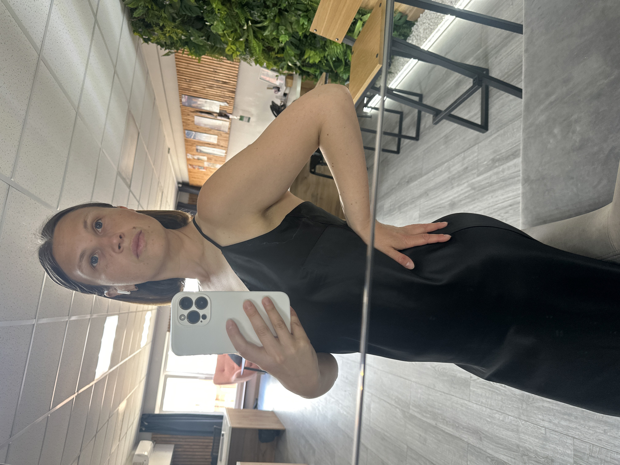 A fair-skinned woman is seen in a mirror selfie indoors. She is wearing a black slip dress and holding a white smartphone. The background includes a ceiling with grid tiles, vertical wood slats with decorative prints, green leafy plants, a black metal structure with a wooden surface, and a partial view of a room with furniture. There is insufficient information to determine whether this image was taken in the city of Chaikovsky, Russia.