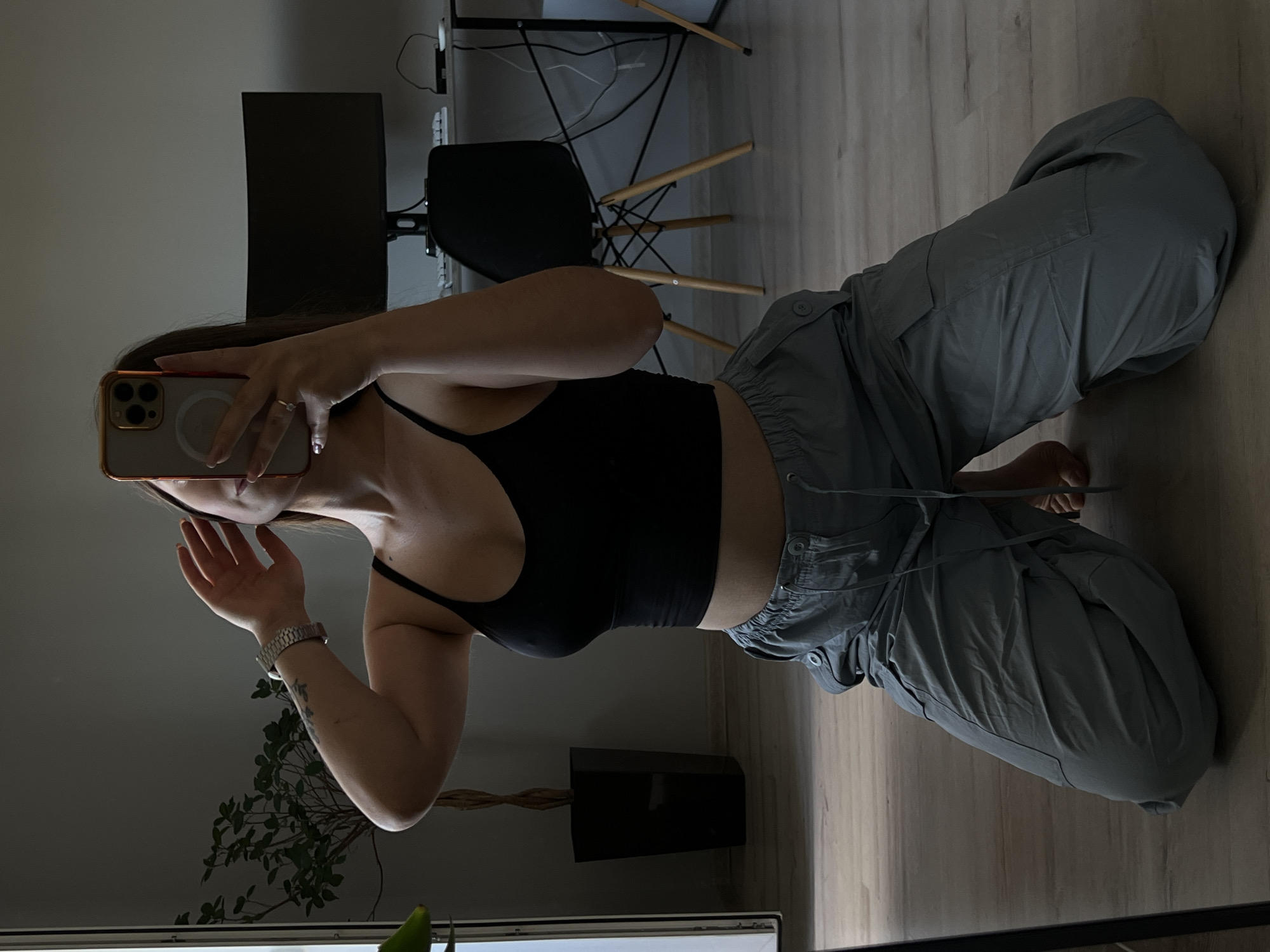 A person is kneeling on a wooden floor, holding a phone to take a mirror selfie. The person is wearing a black crop top and grey cargo pants. In the background, there is a plant in a black pot, a desk with a computer monitor and keyboard, and a black chair. The scene appears to be indoors, likely in an apartment. There is no specific indication that the location is Ternopil, Ukraine.