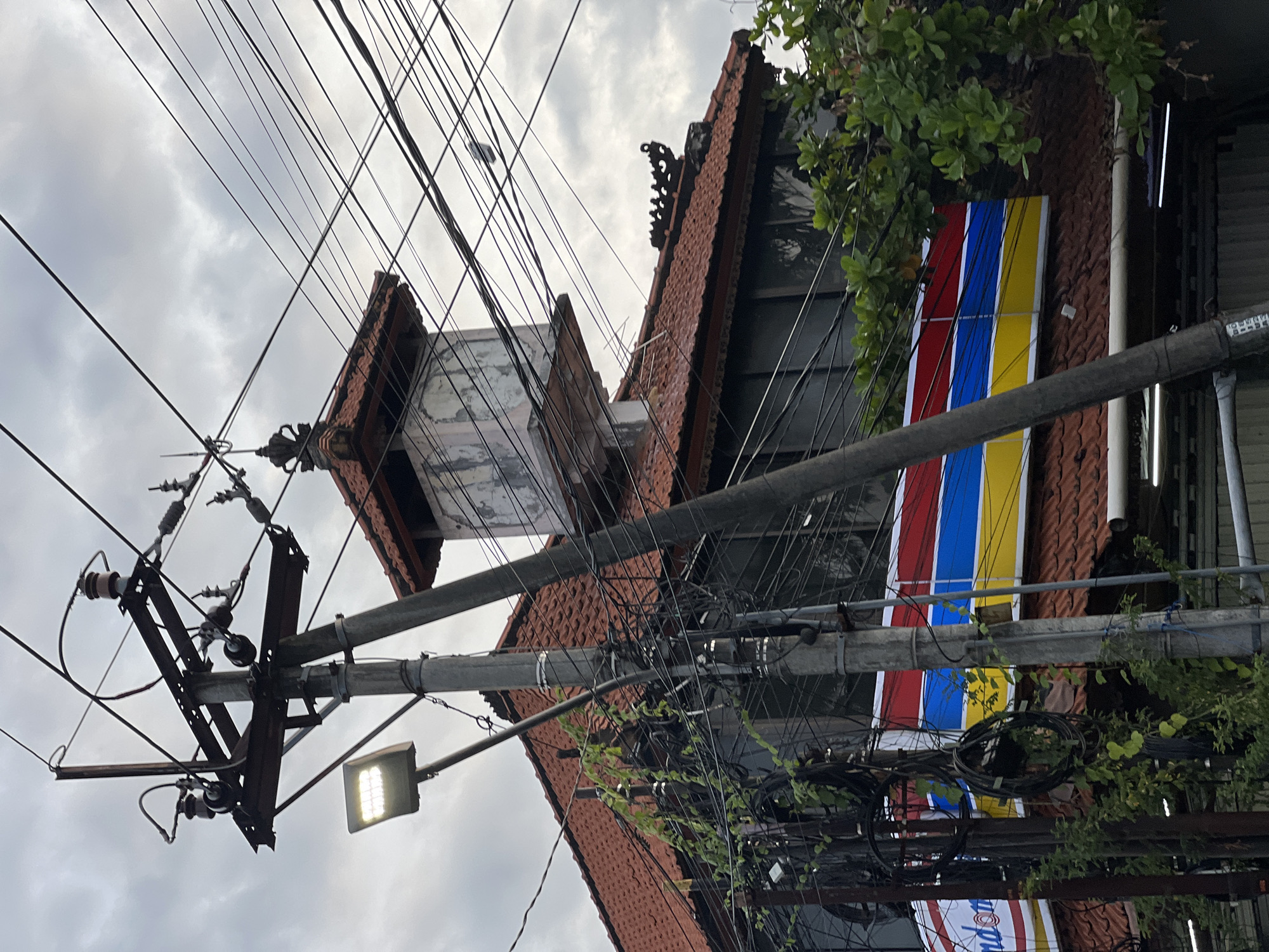 The image captures an urban street scene in Kerobokan Kelod, Indonesia, under an overcast sky. In the foreground, a prominent grey utility pole extends diagonally from the lower left towards the upper right. This pole supports a metal crossarm structure with multiple ceramic insulators and a dense network of black electrical and communication wires. A rectangular LED street light fixture, emitting a bright white light, is attached to this main pole. Numerous black cables are bundled, wrapped, and coiled around the poles, with green leafy vines growing along some of them.
Behind the utility infrastructure, a building with a traditional red-tiled roof is visible. The roof features dark, ornate decorative elements along its ridges and a prominent finial atop a small, elevated, square structure positioned above the main roofline. A large commercial sign is affixed to the building's exterior below the roof, displaying distinct horizontal stripes of red, blue, and yellow, with the partial word "ndom" visible in white lettering on a red background. Green foliage from trees or shrubs partially obscures the right side of the building and some wires. No individuals are present in the scene.