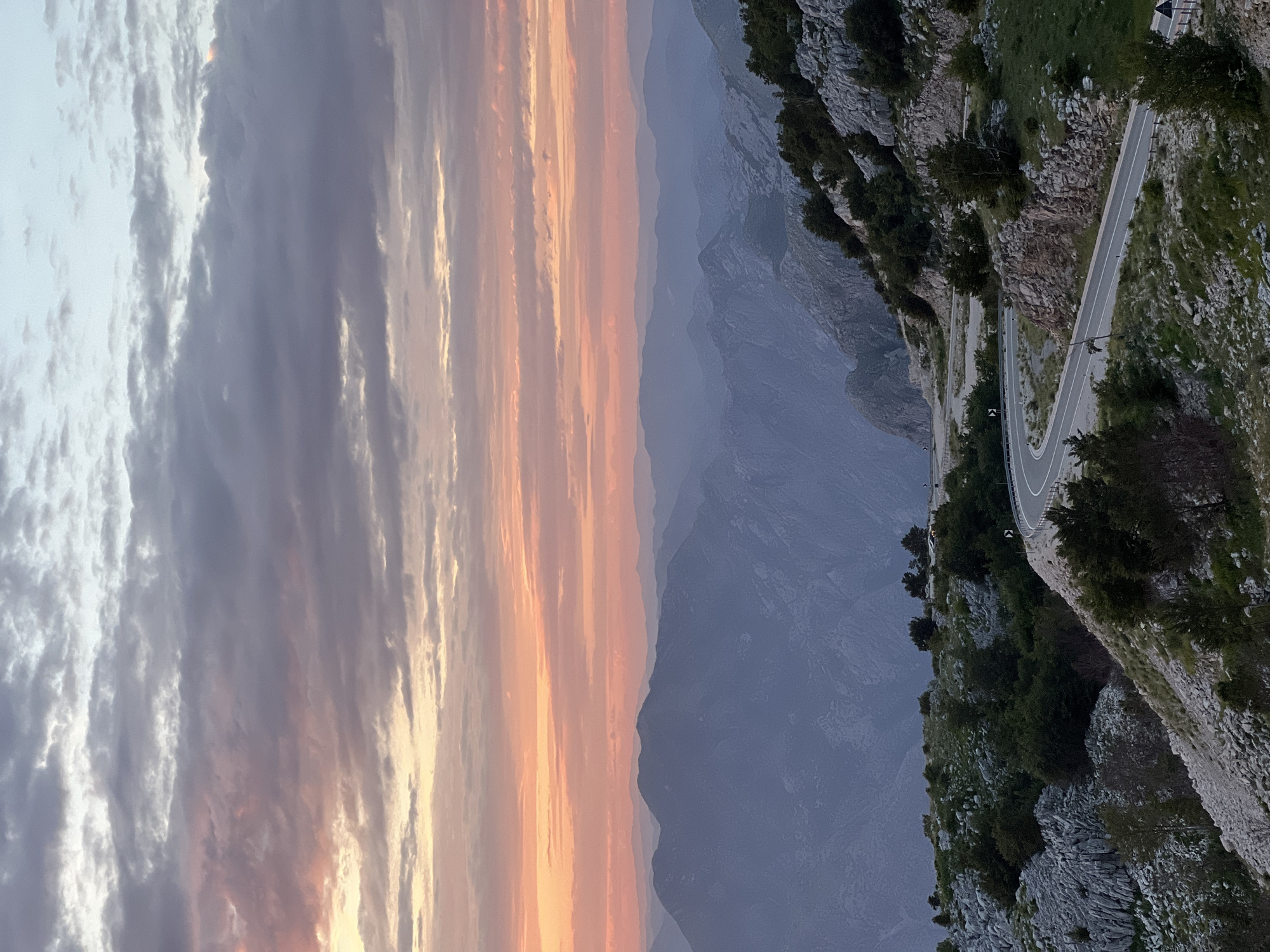 The image depicts a mountainous landscape at either sunrise or sunset, located in Kuk, Montenegro. The sky, occupying the upper portion of the frame, exhibits a gradient from light blue-grey at the top-right to vibrant orange, pink, and red hues along the horizon. Overcast conditions are present, with layered grey and white clouds. Below the horizon line, multiple parallel mountain ranges recede into the distance, displaying atmospheric perspective with progressively lighter, blue-grey tones.
In the midground and foreground, a rugged terrain dominates, characterized by exposed light grey limestone rock formations and sparse green vegetation, including low-lying shrubs and small trees. A two-lane asphalt road with white edge lines and a double solid yellow center line is visible in the lower-right and mid-right sections of the image. The road executes several sharp curves and hairpin turns as it descends or ascends the mountain. Metal guardrails line the outer edges of the road's curves. A black and white chevron-style road sign is positioned on the right side of the road in the midground. A light-colored, possibly white, vehicle is parked on a roadside turnout in the upper right quadrant of the visible road segment. No individuals are present within the scene.