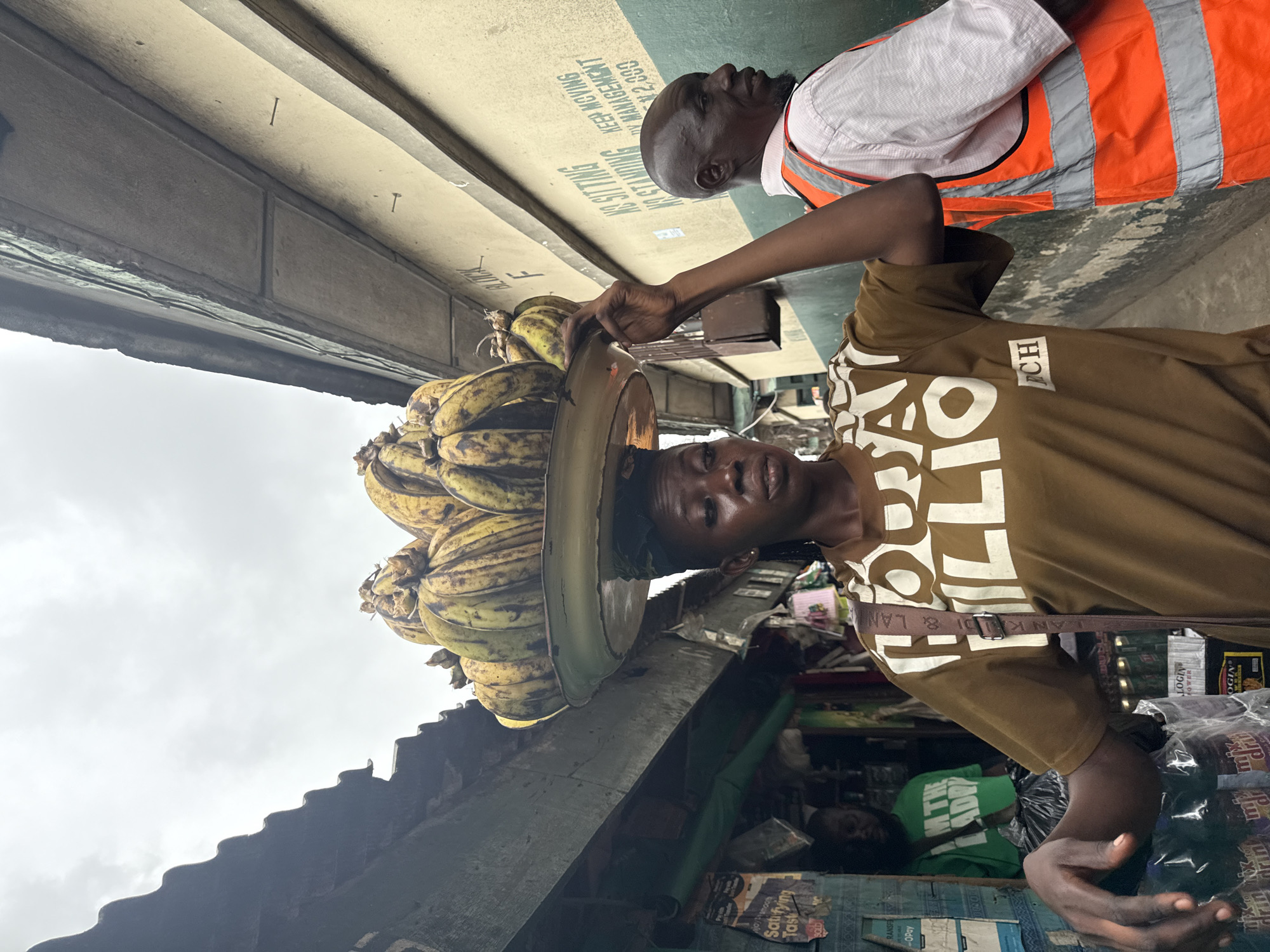 The image captures a street scene in Lagos, Nigeria, with a woman in the foreground balancing a large tray of bananas on her head. She is wearing a brown t-shirt with white text that reads "HOW TO MILIONI". To her right, a man wearing a white shirt and an orange safety vest is visible. In the background, the architecture suggests a bustling urban environment with stalls and buildings lining a narrow street. The sky is overcast, indicating a cloudy day. The overall impression is of daily life and commerce in a Nigerian city.