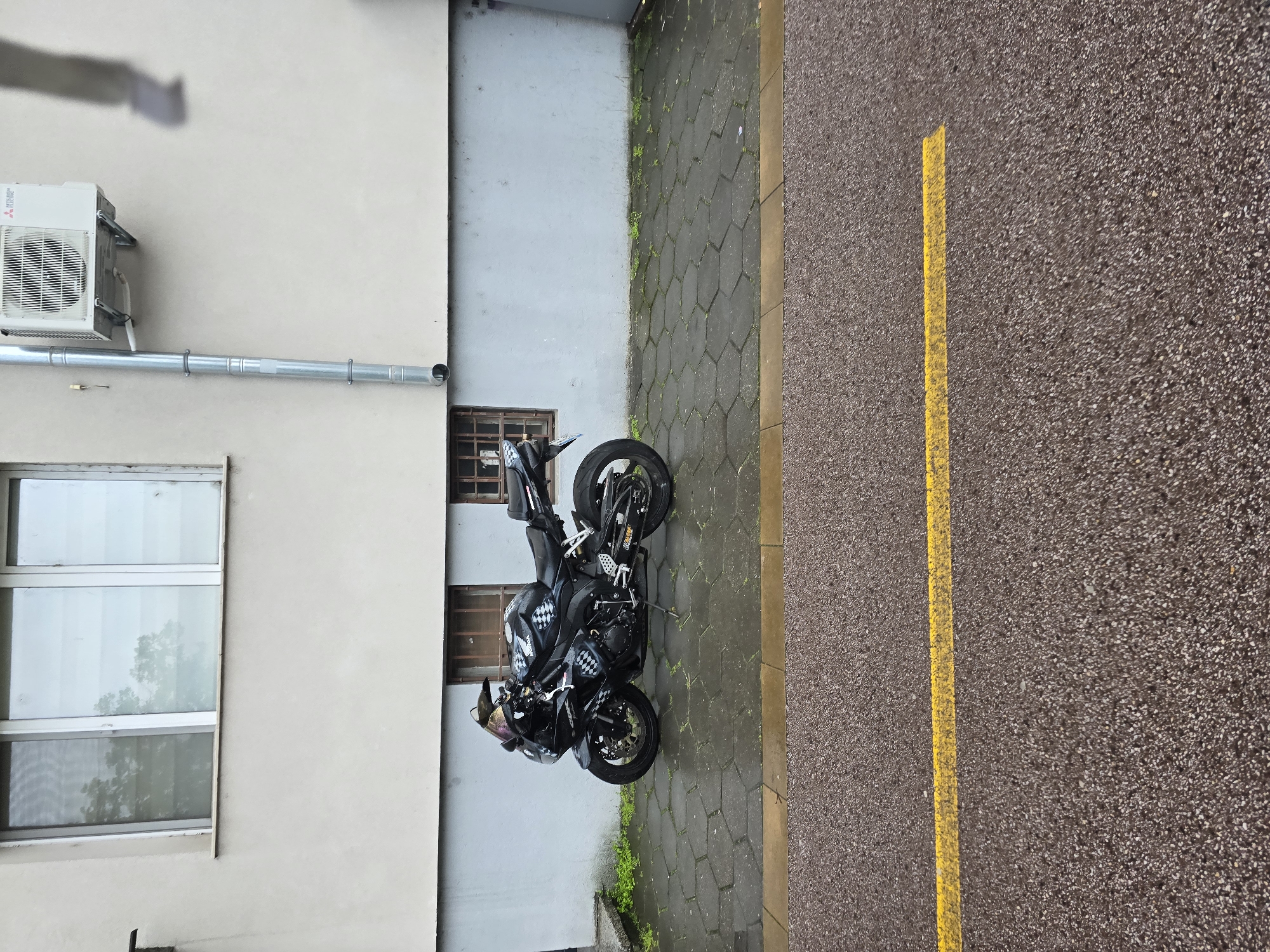 A vertical aerial shot depicts a black motorcycle parked between a building and a road in an unknown city. The motorcycle is positioned on a grey, hexagonal-tiled surface. To the left of the motorcycle is a light-colored building with a window and an air conditioning unit. To the right of the motorcycle is a road with a yellow line.