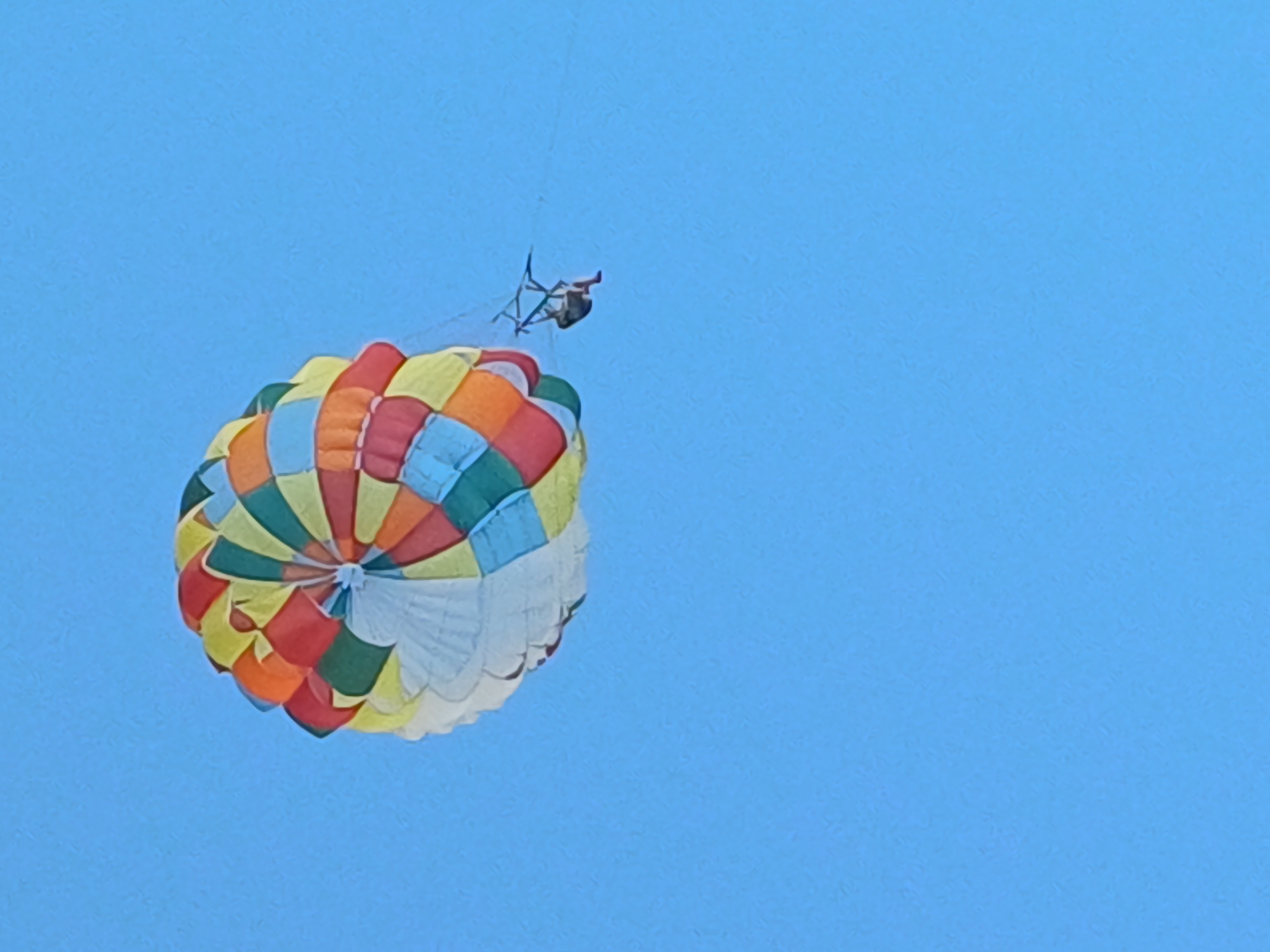 In this media file, a person is parasailing against a clear blue sky. The parasail is a vibrant, multi-colored canopy, featuring segments of red, orange, yellow, green, and blue. A person is seated in a harness beneath the parasail, appearing to be enjoying the activity. The image captures the parasail at an angle, with the person visible above and to the right of its center. The setting is an open sky, suggesting a daytime activity. While no specific location cues are visible in the image itself beyond the sky, the provided context places the scene in Sochi, Russia. The bright sky and the activity of parasailing evoke a sense of recreation and leisure, possibly during fair weather. There is no visible text within the image.