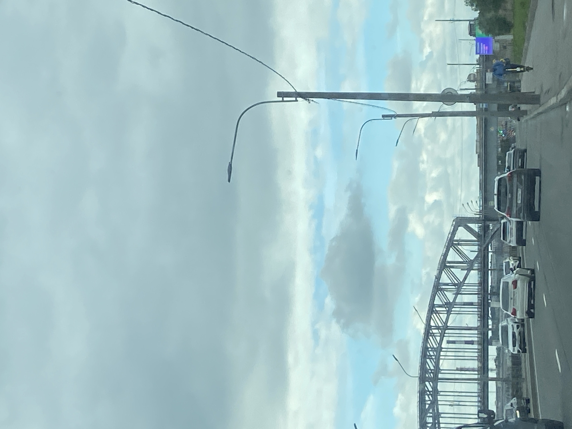The image shows a view from a moving vehicle on a multi-lane highway in Saint Petersburg, Russia, with a large, industrial-style arched bridge in the background. The sky is cloudy, with patches of blue sky visible. There are several cars on the highway, including a white BMW and a white SUV. A person is riding an electric scooter on the right side of the road. Streetlights line the highway. The scene appears to be in an urban or suburban area.