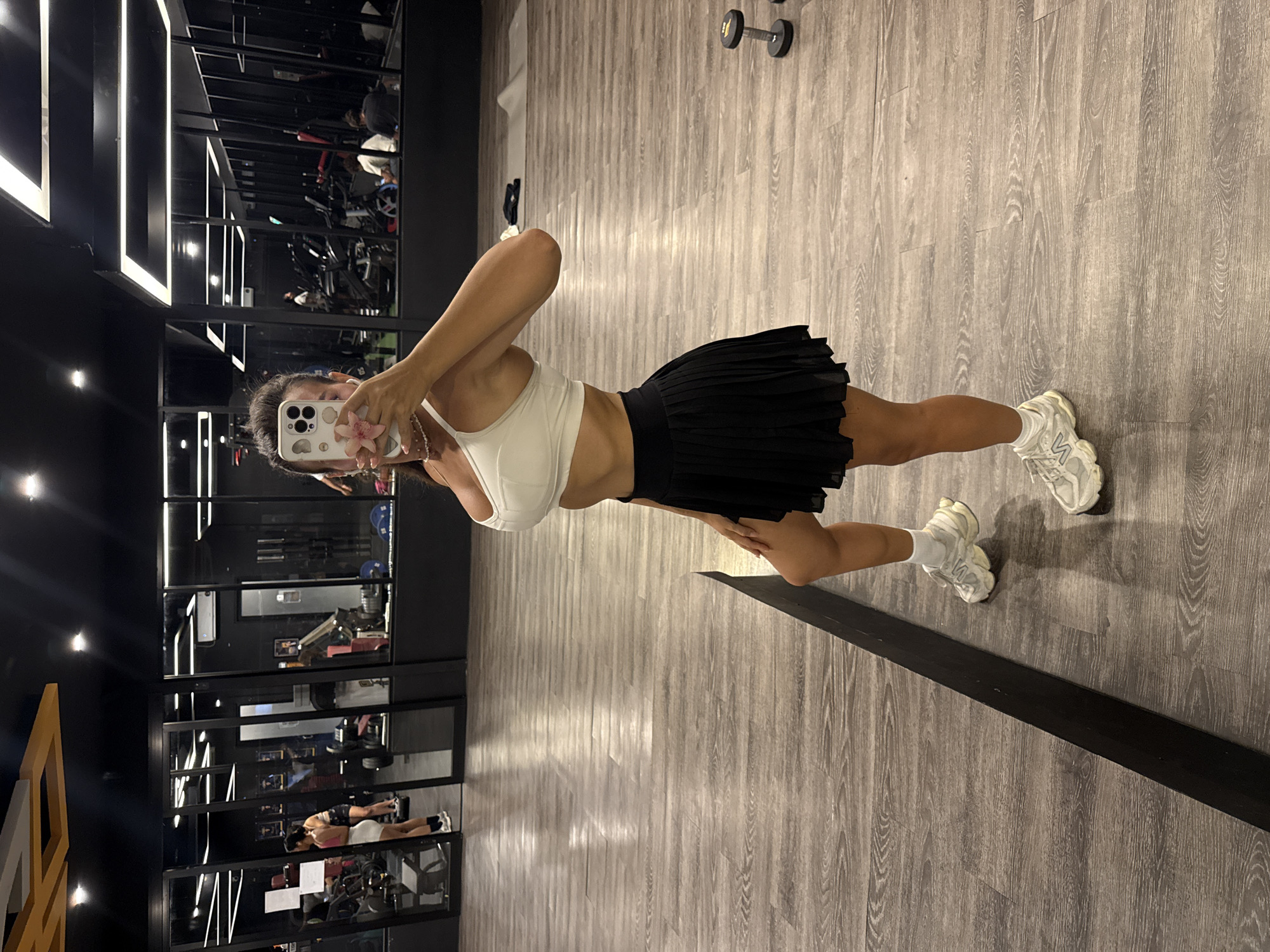 A person is taking a selfie in a gym, as reflected in a mirror. The person is wearing a white sports bra, a black pleated skirt, and white sneakers with white socks. The floor of the gym is gray wood-like panels. There are various exercise machines and weights visible in the background, including treadmills and a dumbbell rack. The gym has black framed mirrors and lighting fixtures. This gym is in North Kuta, Indonesia.