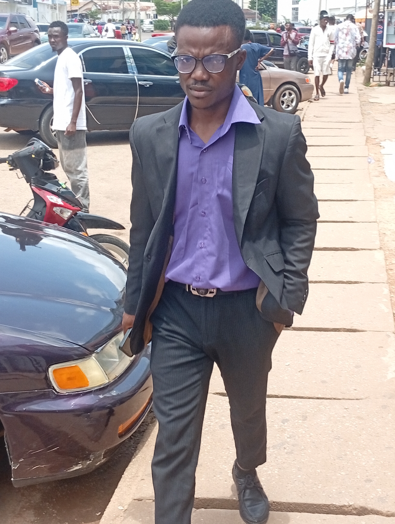 A man wearing a dark suit over a purple dress shirt and dark pants, with black shoes, walks on a paved sidewalk. He is wearing glasses. To his left is a dark-colored car, with part of its front bumper damaged. A red and black motorcycle is parked in front of the car. Behind the man, a person in a white t-shirt and grey pants is standing next to a black car and looking towards the right. Further back, other individuals are walking on the sidewalk and near vehicles. The scene appears to be an outdoor urban environment, likely in Jos, Nigeria, given the context.