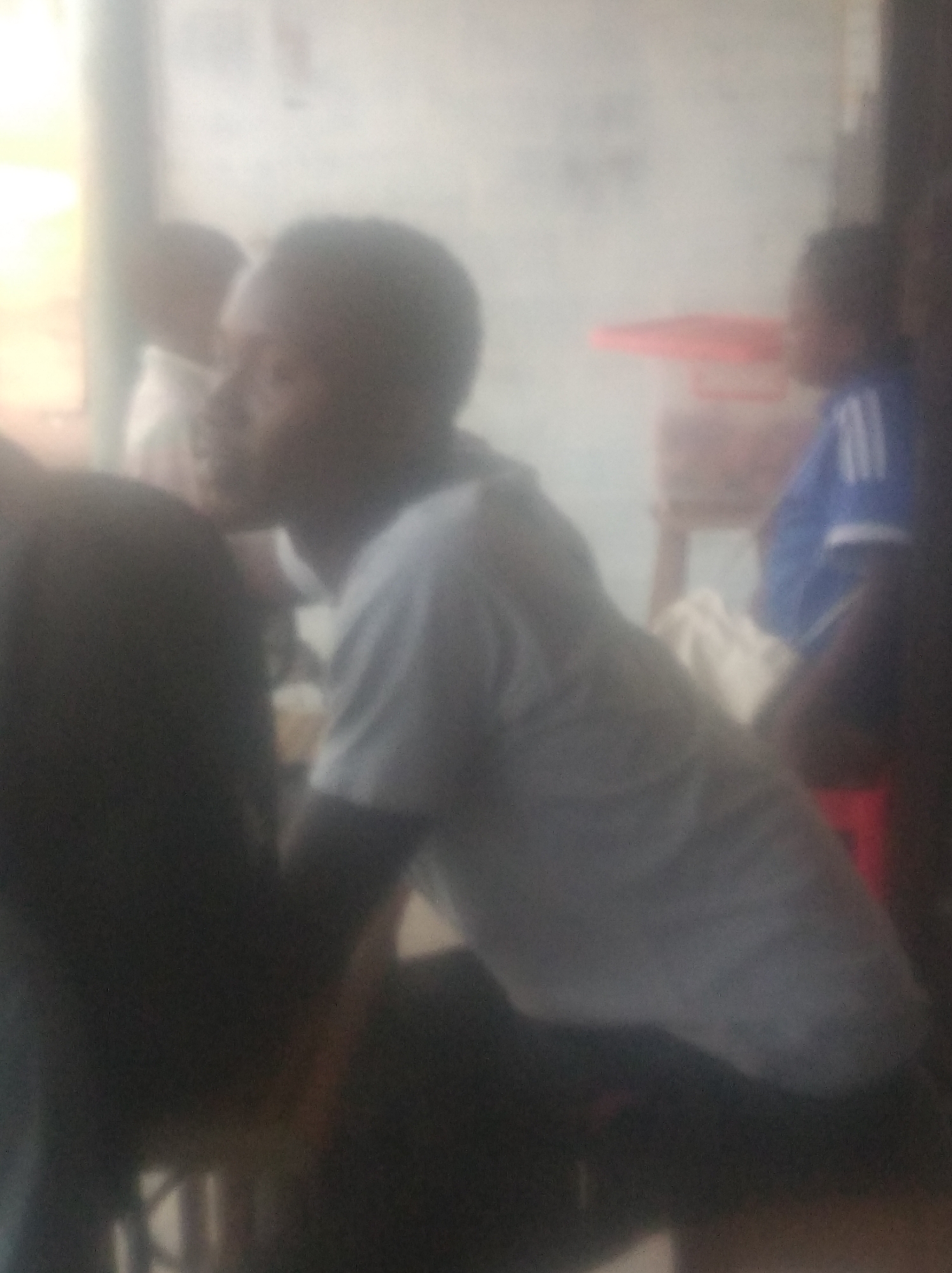 The image depicts an indoor scene, likely in Jalingo, Nigeria, featuring a few individuals in a loosely organized gathering. The lighting is diffuse and appears to be from overhead sources, casting a soft glow on the subjects. The primary subject in the foreground is a young Black male wearing a light-colored t-shirt. He is positioned in profile, facing towards the right of the frame, with his head tilted downwards as if engaged in conversation or observing something.
Behind him, partially obscured, is another individual, also a Black male, wearing a blue t-shirt with white stripes on the sleeves. This individual appears to be standing or leaning. To the left of the main subject, a dark silhouette of a person is visible, their form indistinct. The background suggests an interior space with a light-colored wall, possibly with some faint markings or textures. A red object, possibly a piece of furniture or a container, is visible in the mid-background. The overall impression is one of a casual, informal social interaction within a somewhat dimly lit indoor environment.