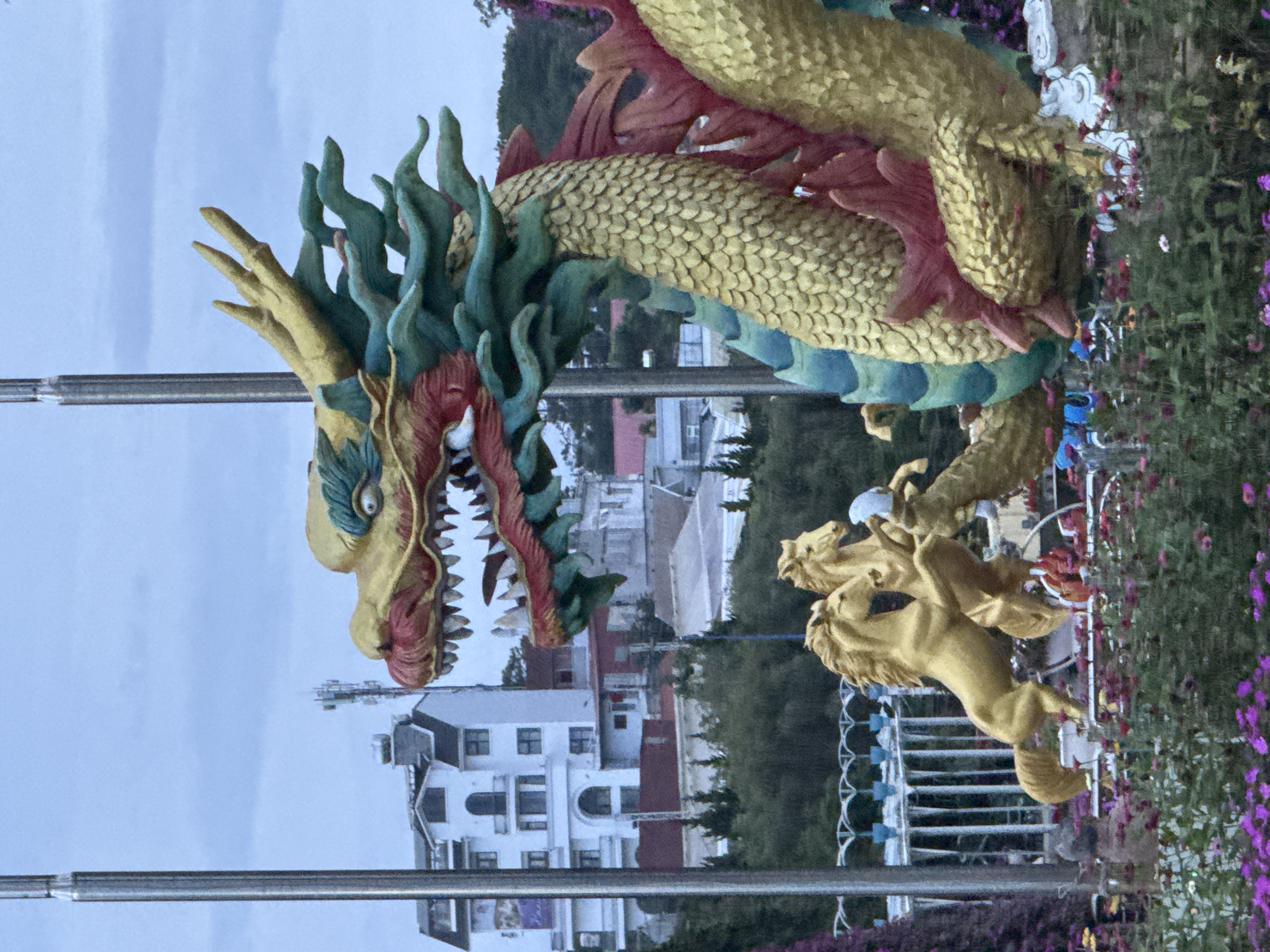The image shows a large, ornate statue of a golden dragon with green and red accents. The dragon's head is prominently featured in the foreground, with its mouth open revealing sharp teeth. Behind the dragon, there are two golden horse statues rearing up, and a white fence. In the background, there are buildings, trees, and a telecommunications tower. The scene appears to be a park or garden in Lâm Viên - Đà Lạt, Vietnam, given the style of the statues and the architecture in the background. The sky is overcast.