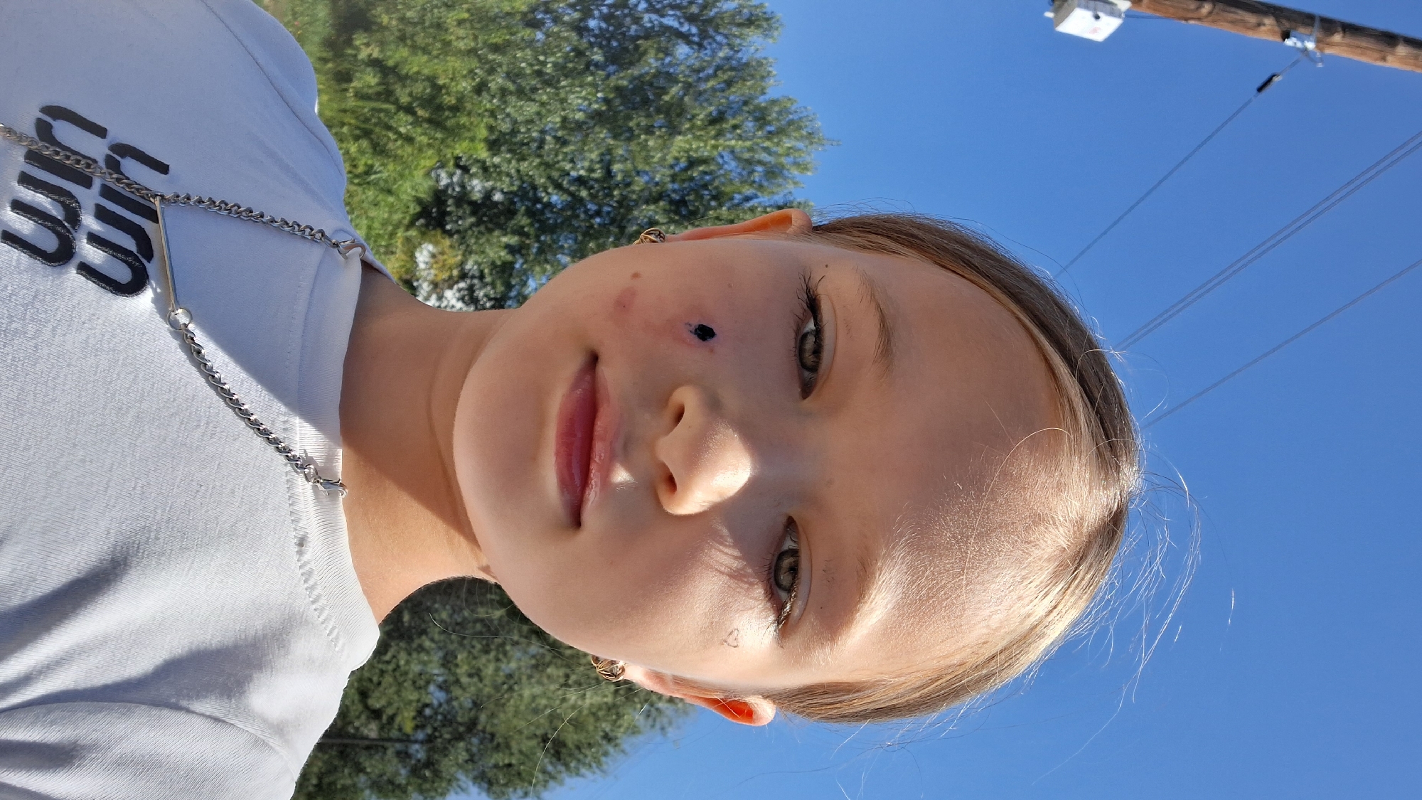 A young girl is featured in a close-up, sideways selfie. She has light brown hair and her eyes are a striking green. Her lips are slightly parted, revealing a hint of pink. A small, dark mark is visible on her cheek, near her eye, and another faint heart shape is drawn near her other eye. She is wearing a white t-shirt with a silver chain draped across it. The chain has a clasp and a small bar charm. The background shows a clear, bright blue sky, some green foliage from trees, and a wooden utility pole with wires. The lighting suggests it is daytime, likely sunny, and outdoors. The girl's expression is neutral or perhaps a slight hint of a smile. No visible text is discernible on the girl's clothing.