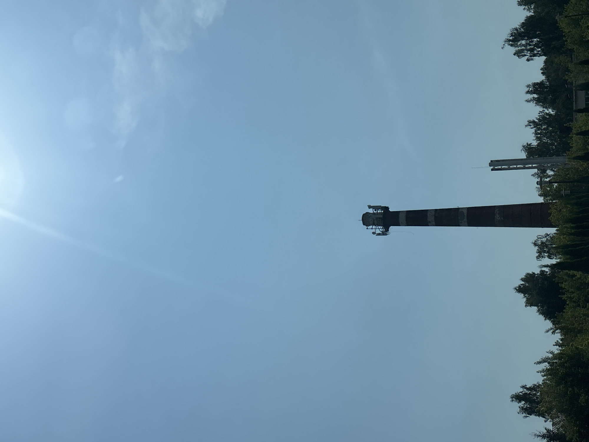 The image depicts a tall, red and white striped brick chimney against a clear blue sky. The chimney has equipment attached to its top. In the background, a line of dense green trees and foliage forms the lower edge of the image. To the right of the chimney, a slender, grey, modern tower with antennas is visible. The overall scene is outdoors under bright daylight, with a lens flare visible in the upper right portion of the sky. The location is identified as Zvenigorod, Russia.