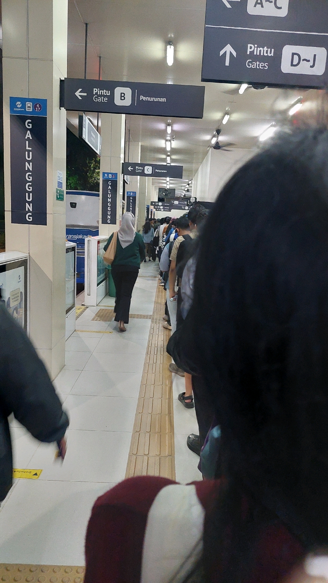 The image captures a scene at the Transjakarta Corridor 13, Lebak Bulus Terminal, South Jakarta, Indonesia, likely during a period of moderate passenger activity. In the foreground, a person's back is visible, obscuring much of the view. The midground features a line of passengers queuing, mostly facing away from the camera and towards the direction of travel. A woman in a green hijab and dark pants is walking away from the camera, heading towards the passenger gate. She carries a light brown tote bag.
Overhead, directional signs are prominently displayed. One sign indicates "Pintu B Penurunan" (Gate B Descent) with an arrow pointing left, suggesting an exit or lower level access. Another sign points upwards with "Pintu Gates D~J", indicating a different gate section. Further down the corridor, a similar sign for "Pintu C Penurunan" (Gate C Descent) is visible with a leftward arrow. Vertical blue signs with white text identify "GALUNGGUNG" which is the name of one of the stations on Corridor 13.
The environment appears to be an elevated busway station. The floor is tiled, with textured yellow tactile paving strips along the edges of the walkway and near the platform. Fluorescent lighting fixtures are mounted on the ceiling, along with several ceiling fans, suggesting a climate-controlled or ventilated indoor space. The overall atmosphere is utilitarian, typical of public transportation hubs.