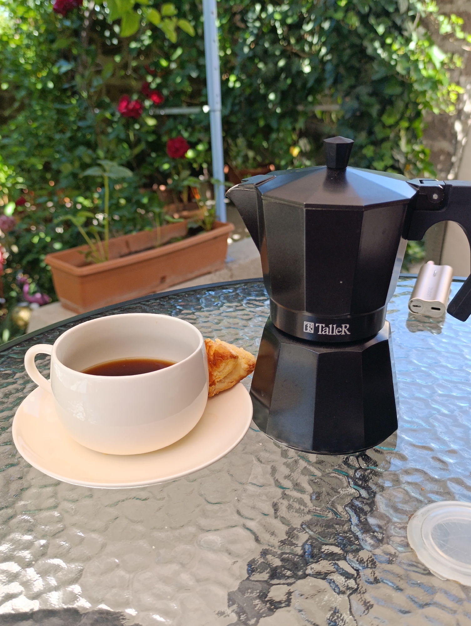 An overhead, eye-level medium shot shows a coffee set on a glass table in an outdoor garden setting. A black espresso maker with a silver accent is on the right side of the frame, and a white cup of dark coffee with a croissant is on the left. The table's textured glass surface reflects the surrounding greenery and sunlight. Behind the table, a blurred background of lush green foliage and red roses suggests a garden environment. The lighting is bright and natural, indicating it is daytime.
