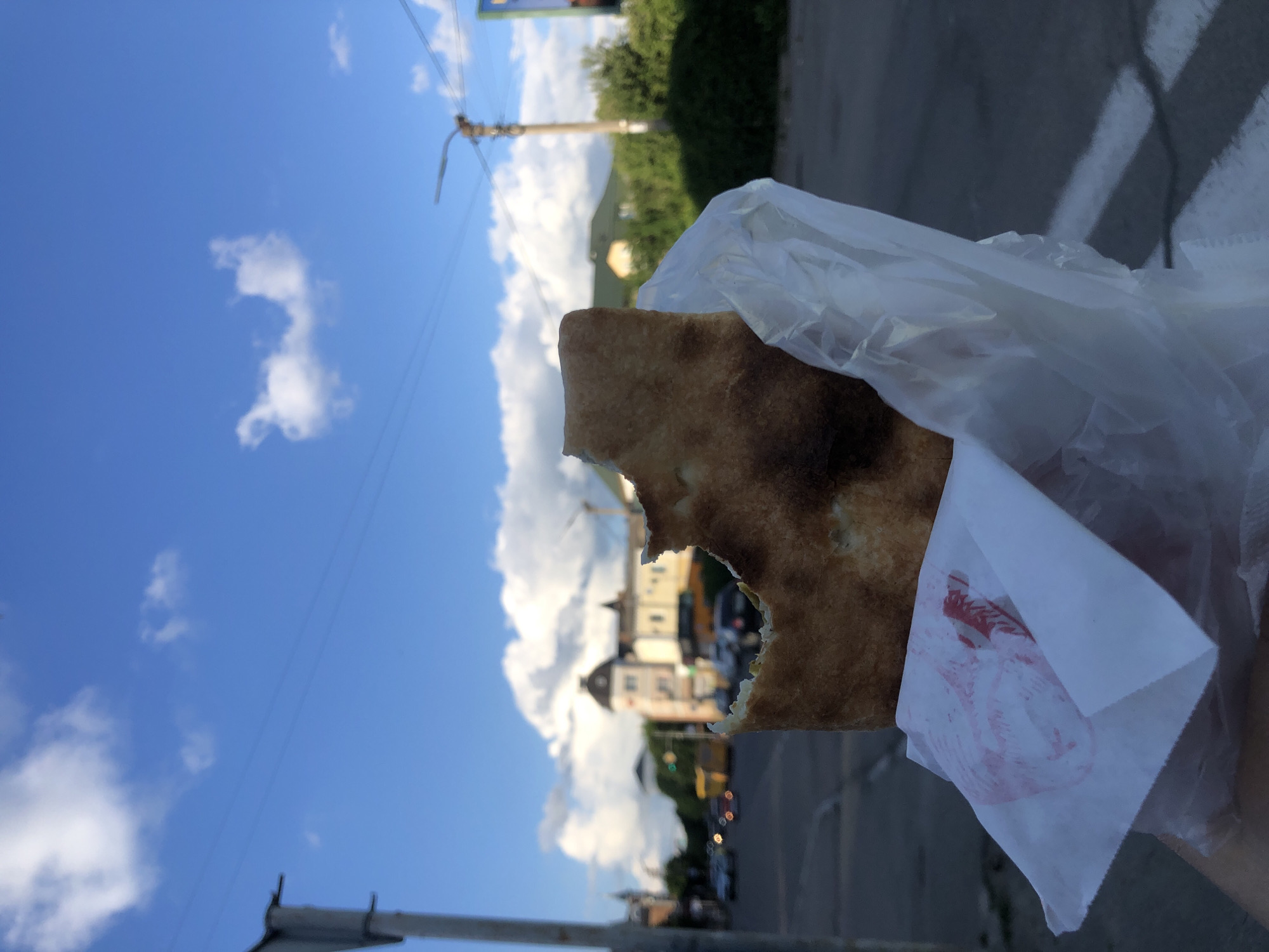The image displays a close-up of a partially eaten piece of flatbread, possibly lavash, held by an unseen hand. The flatbread is golden-brown with some darker, baked spots, and a clear bite mark reveals a lighter interior. It is held within a translucent plastic bag, which is further wrapped in white paper.
The background shows a bright, sunny outdoor scene. The upper portion of the frame features a clear blue sky with several scattered white cumulus clouds. Below the sky, a street or road is visible, flanked by trees and buildings in the distance. A utility pole with power lines extends into the sky from the right side. Distant buildings, one with a distinctive rounded or dome-like roof, are visible but appear slightly out of focus. Blurred vehicles can be seen on the road.
The time of day appears to be daytime, likely afternoon, given the bright illumination and visible shadows. The weather is clear and sunny. On the white paper wrapper, a red illustration is visible, resembling a stylized bird, possibly a chicken or rooster, but no discernible text is present. The overall setting suggests an urban or semi-urban environment where someone is enjoying a snack outdoors.
